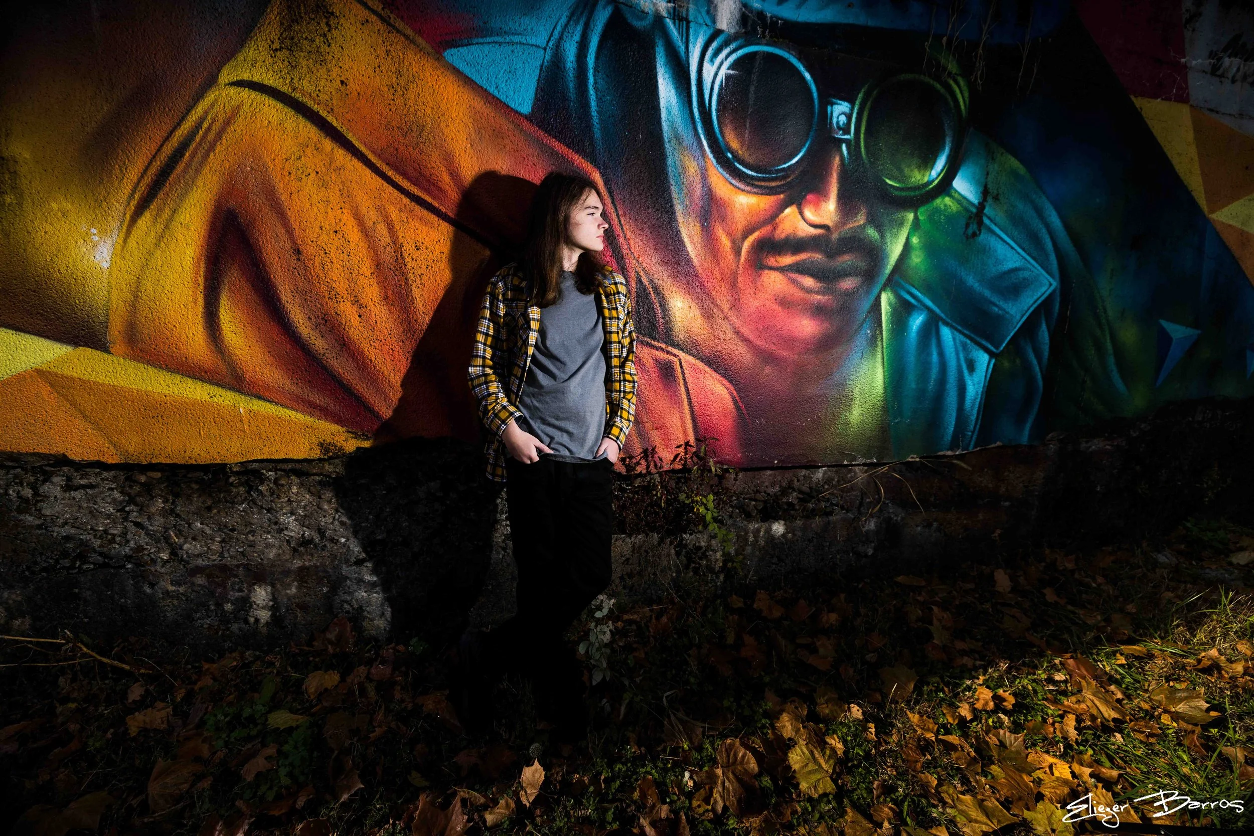 A young woman stands in front of a colorful mural featuring a man's face with large goggles. She is wearing a gray t-shirt, a yellow and black plaid shirt, and black pants, and has her hands in her pockets. Nighttime scene, with fallen leaves on the 