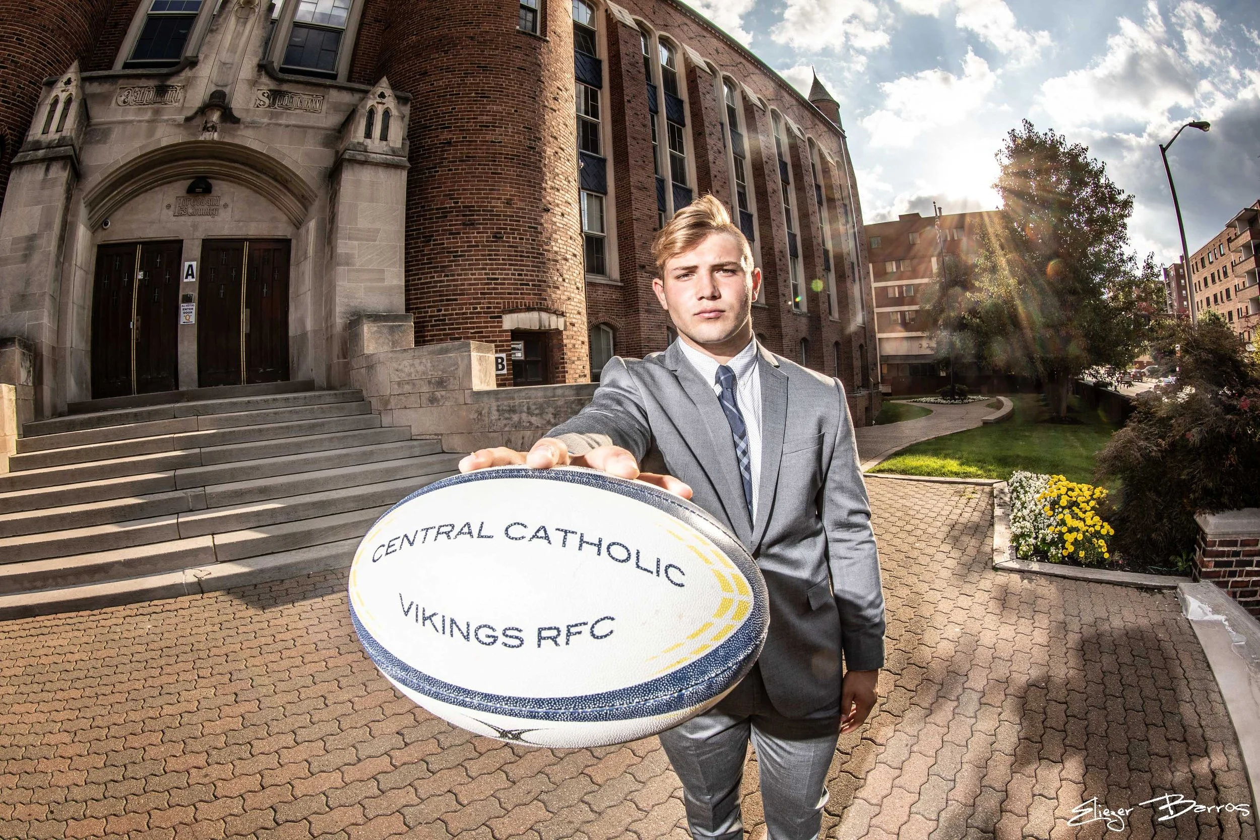 Senior Pictures Pittsburgh at Central Catholic High School. Young man in a gray suit holding a rugby ball in front of the historic school building for academic and athletic senior pictures.