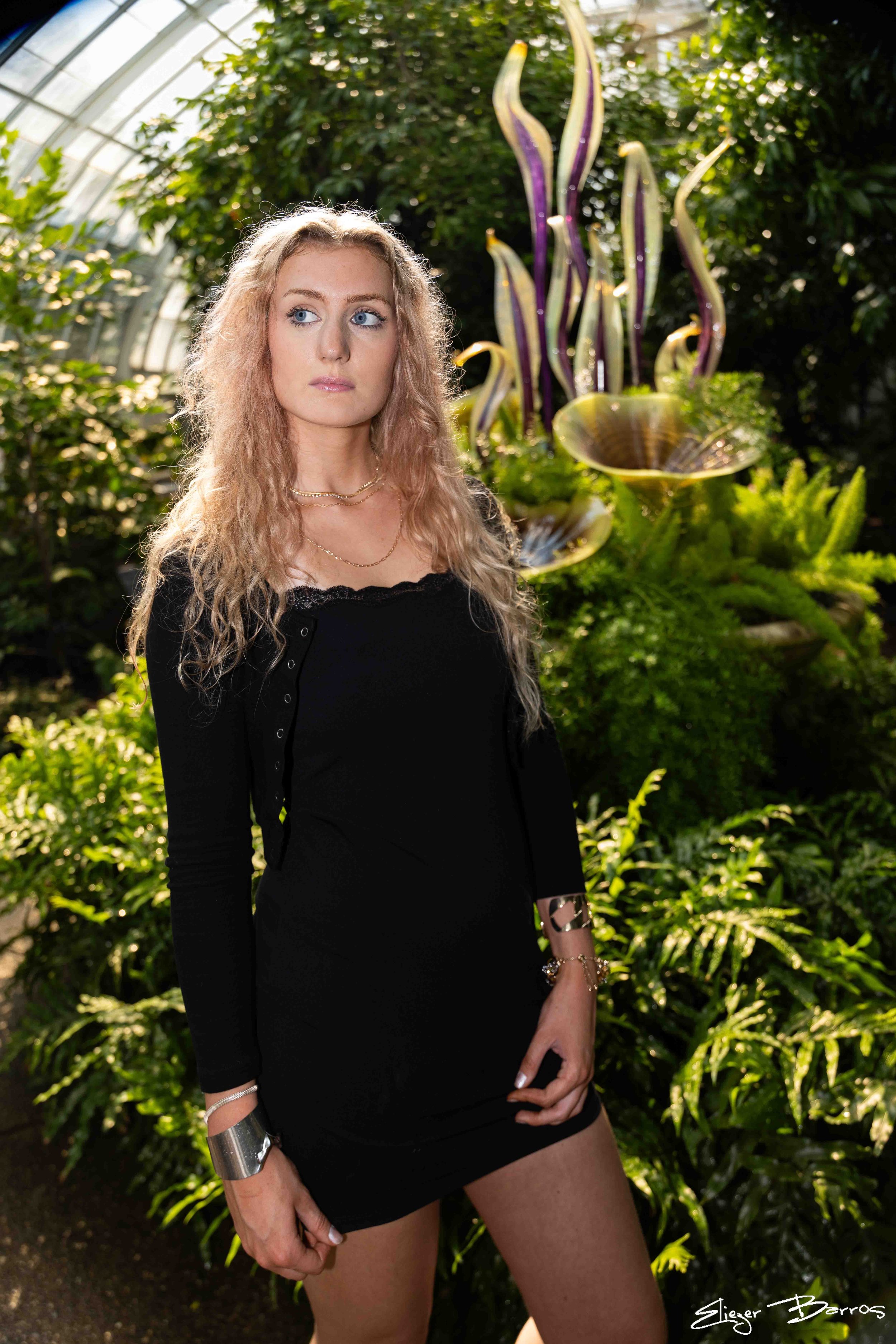 A woman with long, wavy blonde hair wearing a black dress with laced neckline, silver jewelry including necklaces, bracelets, and a cuff, standing in a lush, green conservatory or garden with exotic plants and tall, purple and yellow striped flowers 