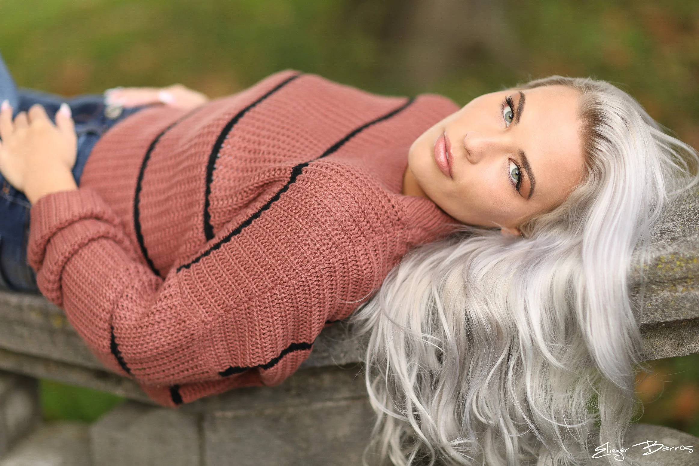 A woman with long, silver hair lying on a stone surface outdoors, wearing a pink and black knitted sweater and dark pants, with a blurred natural background.