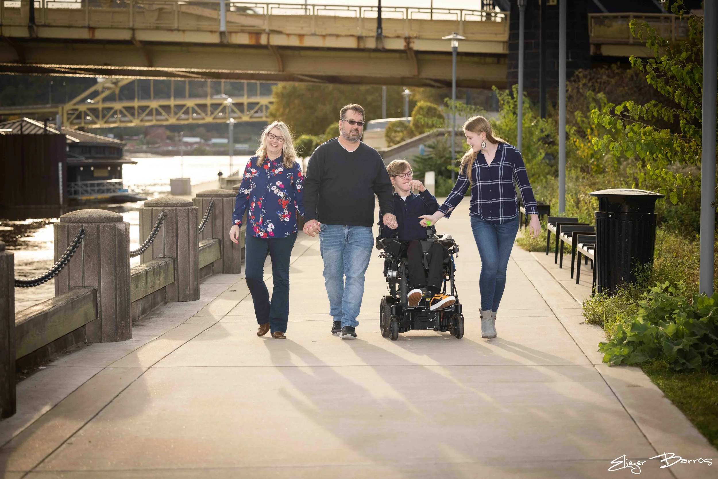 Four people, family or friends, walking along a riverside walkway in the late afternoon or early evening. One person uses a motorized wheelchair, and another is holding their hand. They are smiling and enjoying each other's company. The background fe
