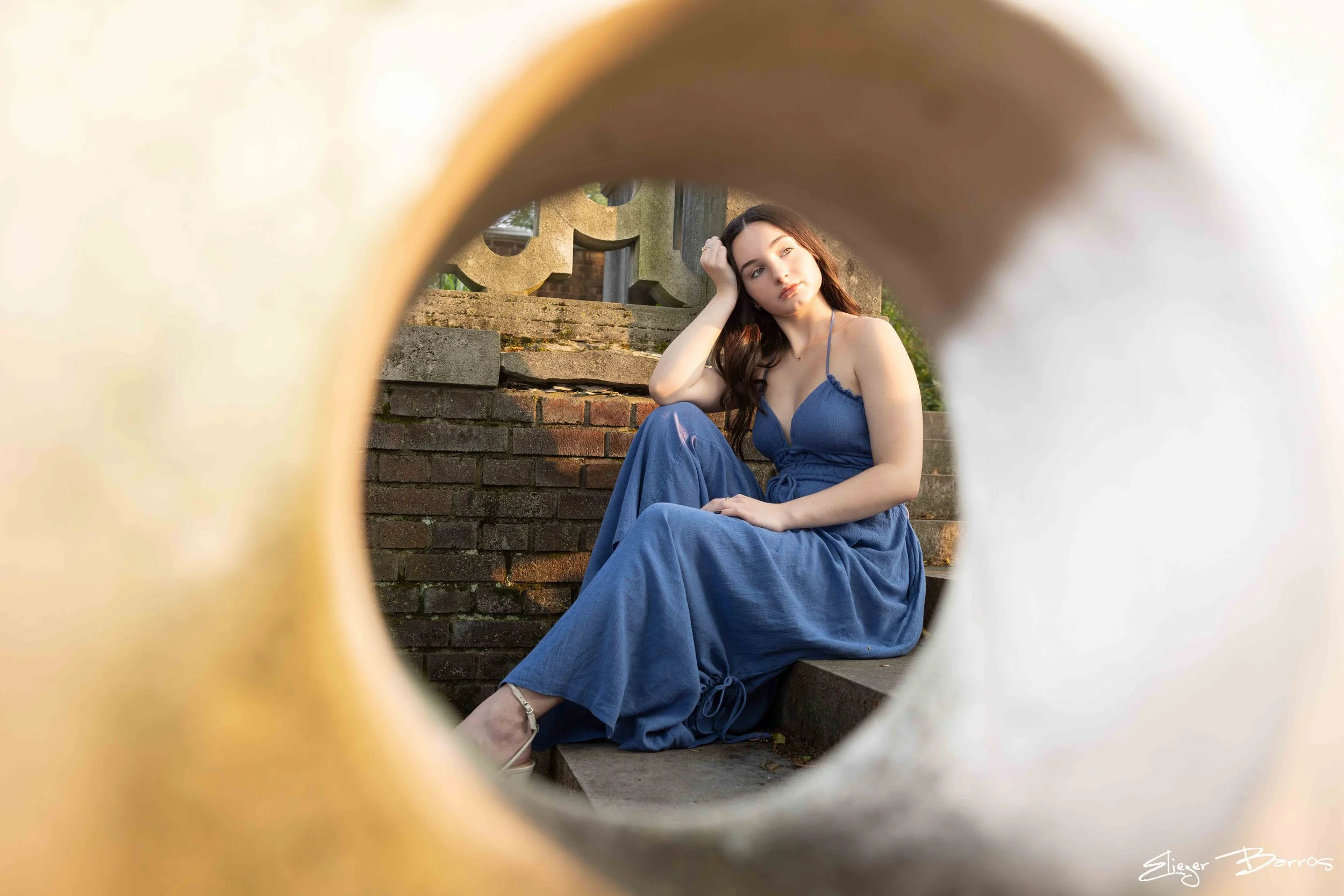 Young woman with long brown hair, blue dress, sitting on steps outdoors with her hand on her head, seen through a circular frame.