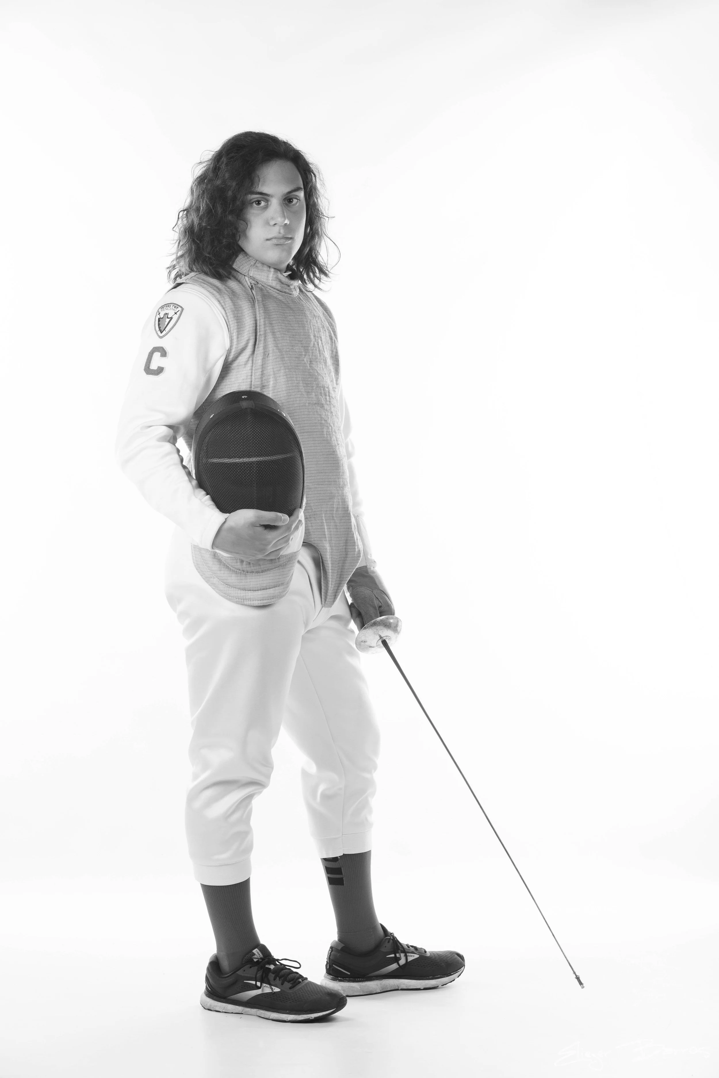 A person dressed in fencing gear holding a fencing foil, standing in a studio with a plain background.