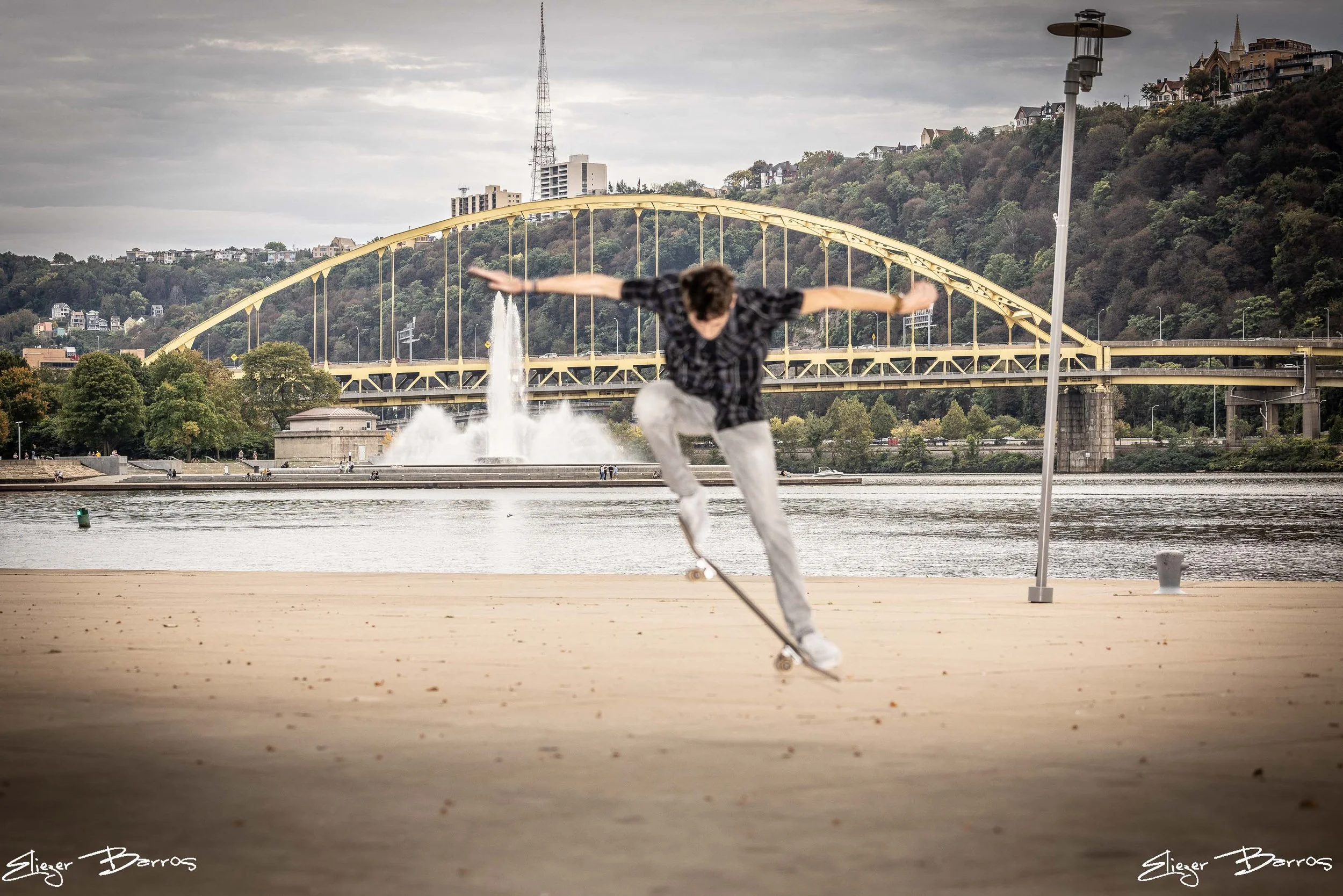 A skateboarder in a black plaid shirt skateboarding on a pier near the water with a bridge and hillside with houses in the background.