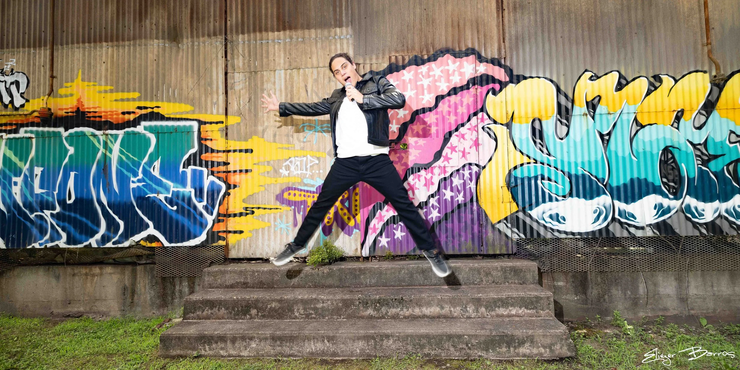 A person jumping in front of colorful graffiti art on a corrugated metal wall, holding a microphone, wearing a leather jacket, white shirt, black pants, and sneakers.