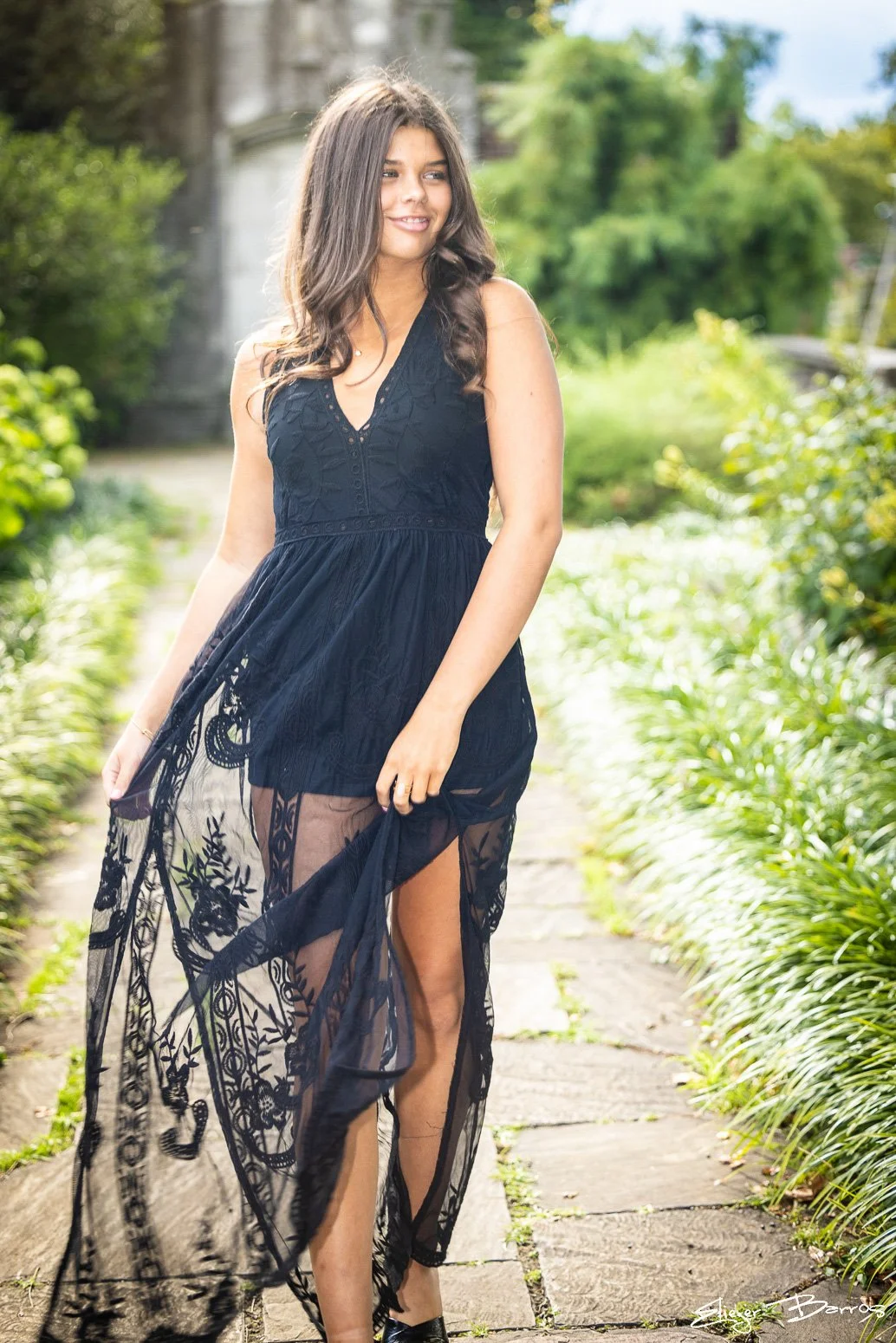A woman in a black lace dress with a sheer skirt standing on a stone pathway in a lush outdoor garden.