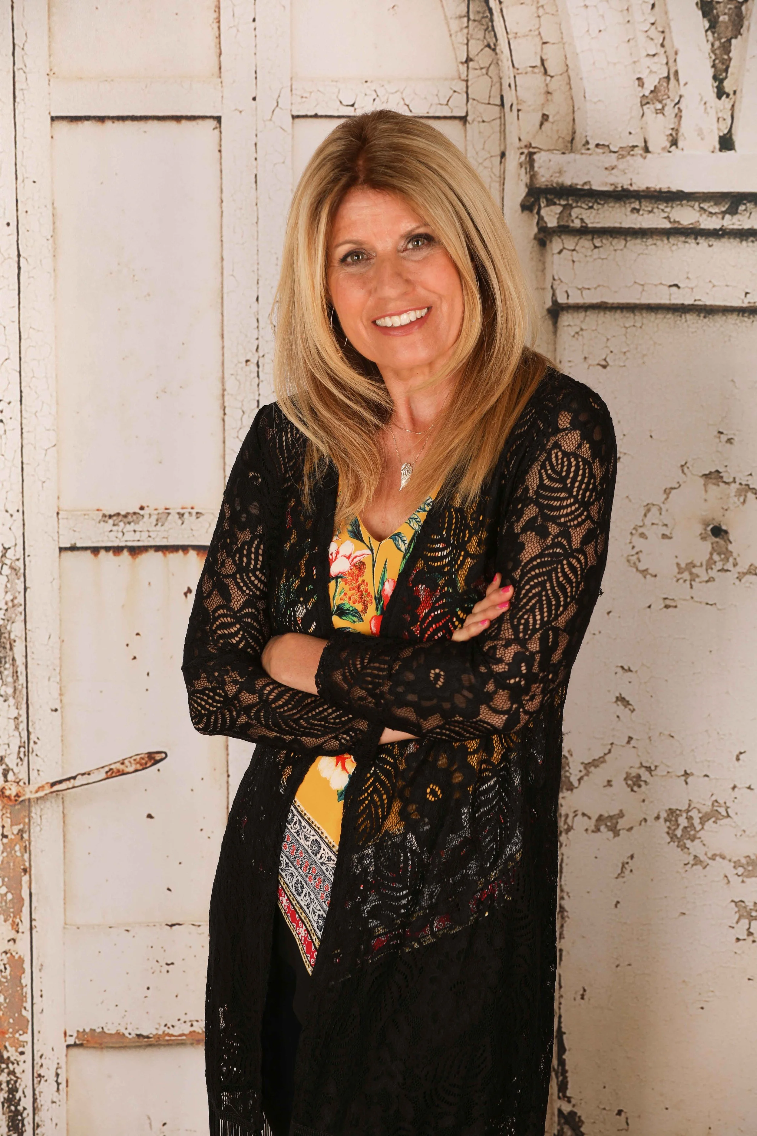 A woman with blonde hair smiling and crossing her arms, wearing a black lace cardigan over a colorful floral dress, standing in front of a distressed white wooden door.