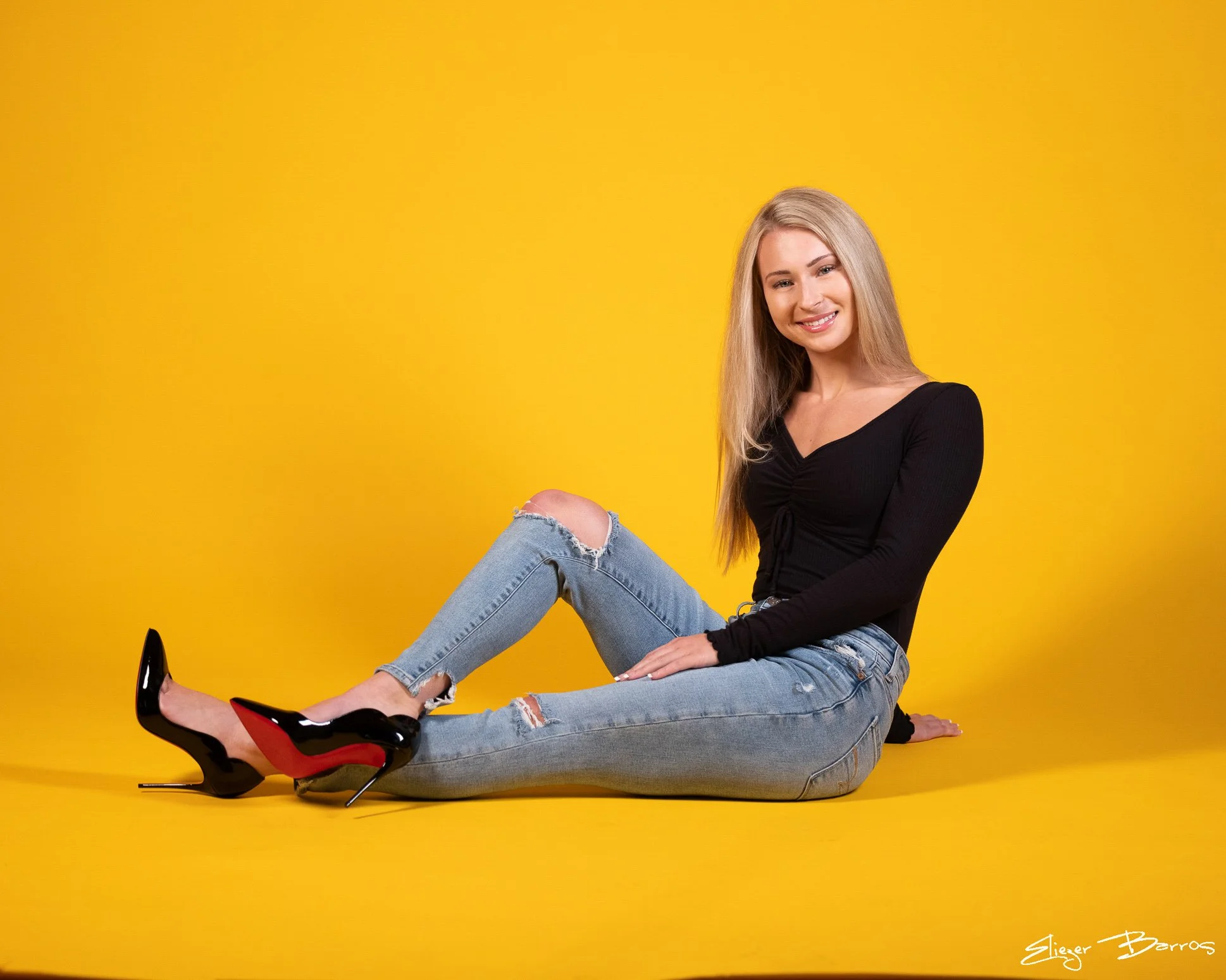 A young woman with long blonde hair sitting on the floor against a bright yellow wall, smiling, wearing a black long sleeve top, ripped jeans, and black high heels with red soles.