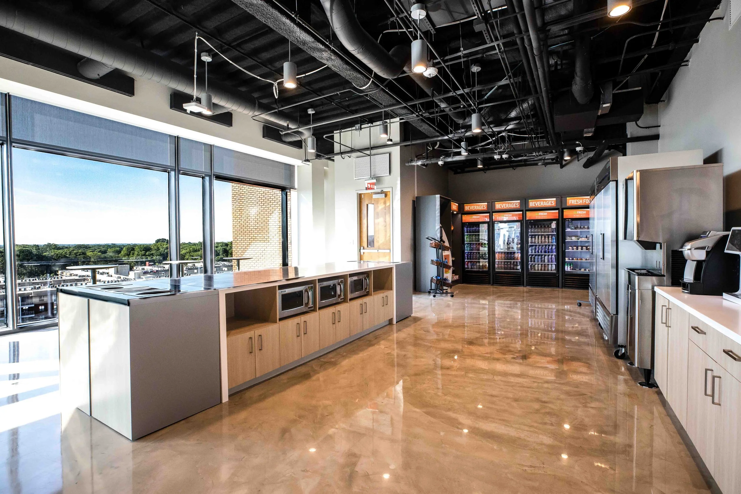 Modern office break room with large windows, a counter with microwaves, beverage refrigerators, and a coffee machine, overlooking a cityscape with trees.