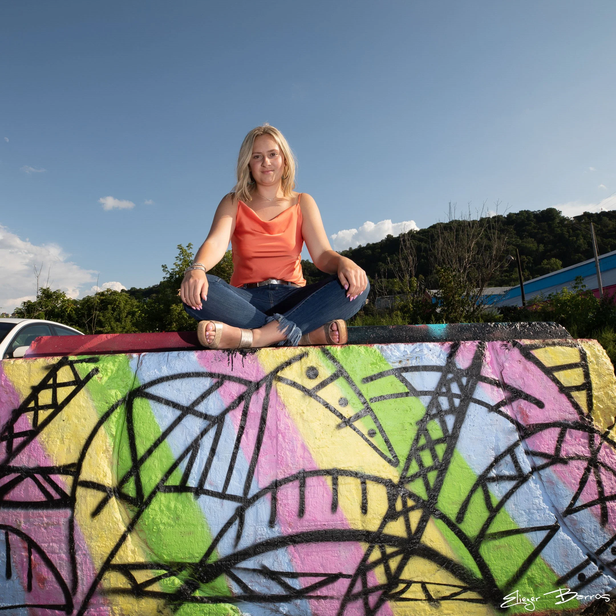 A woman with blonde hair sitting cross-legged on a colorful, graffiti-covered concrete surface outdoors on a bright, clear day with a blue sky and trees in the background.