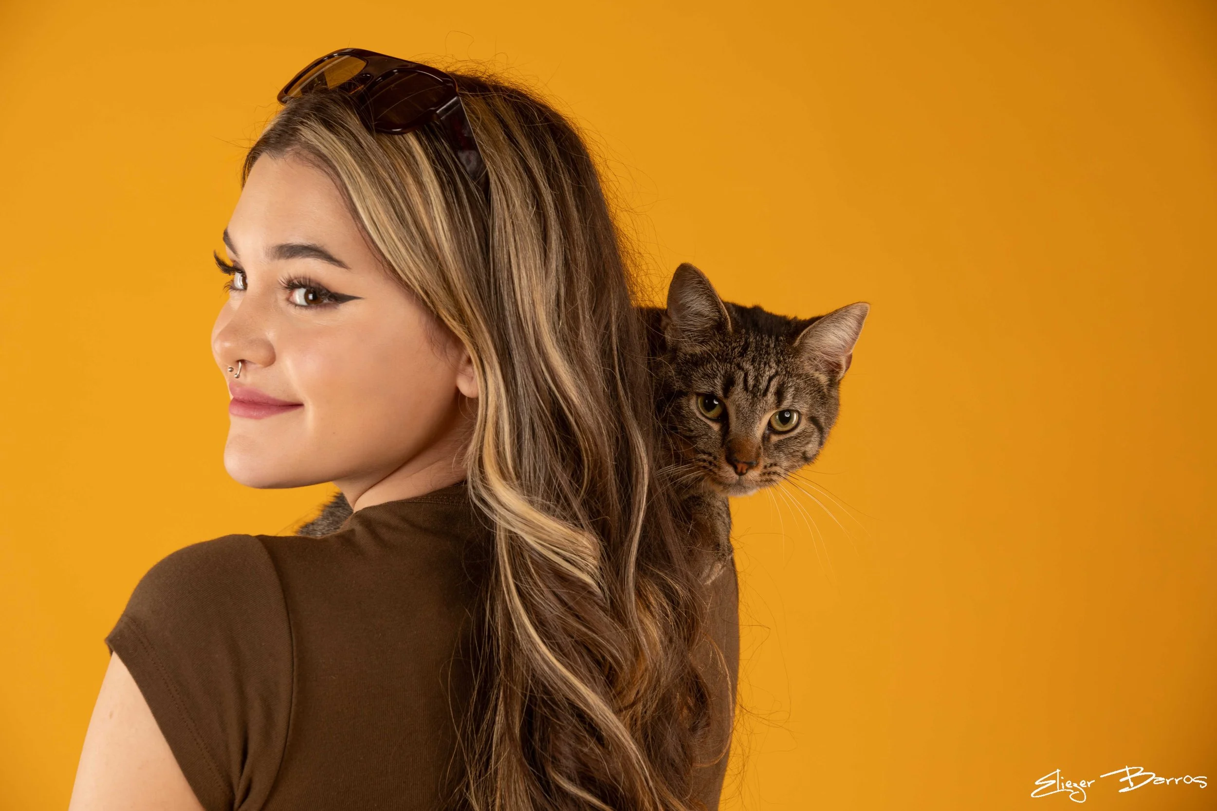 Young woman with long wavy brown hair, wearing sunglasses on her head, looking to her left, with a tabby cat on her shoulder against a solid yellow background.