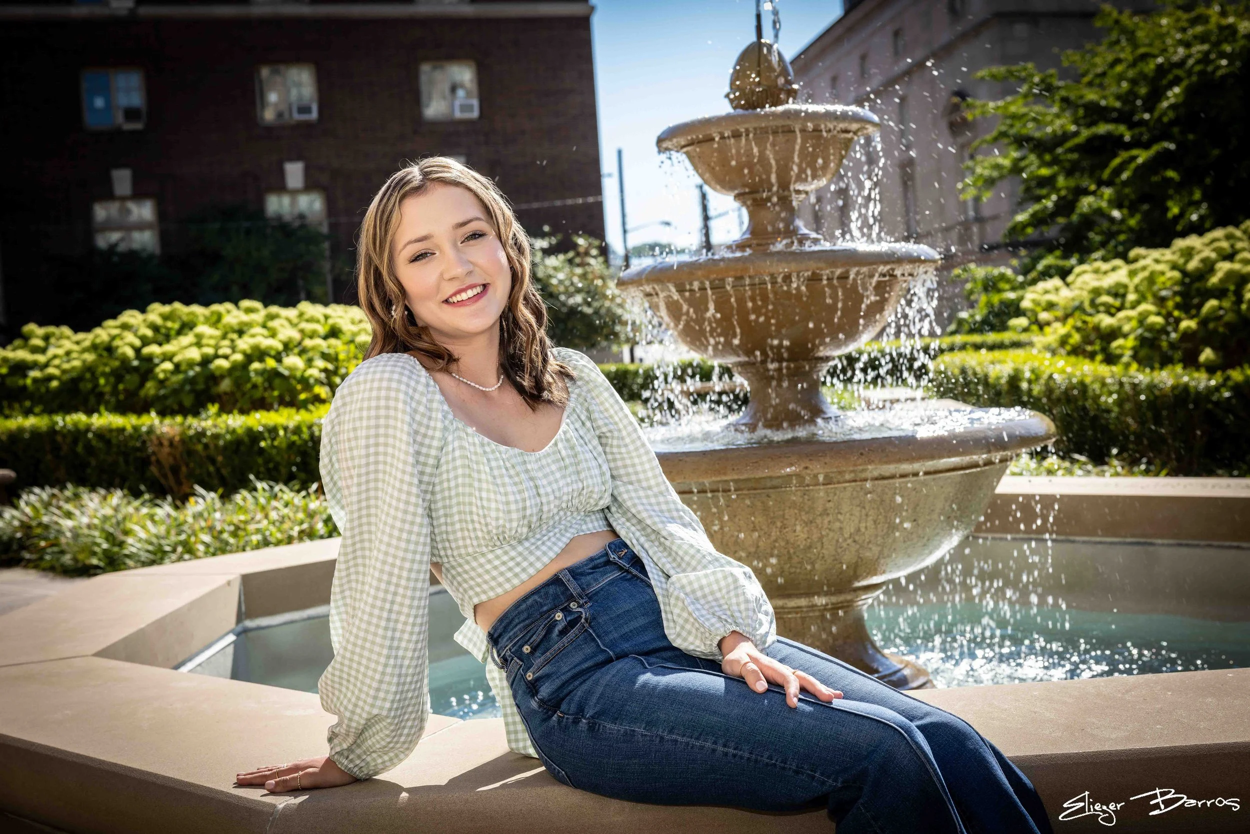A young woman with wavy light brown hair, wearing a green and white checkered blouse and blue jeans, sitting on a ledge beside a multi-tiered fountain with water pouring from the top, in an urban park with greenery and brick buildings in the backgrou