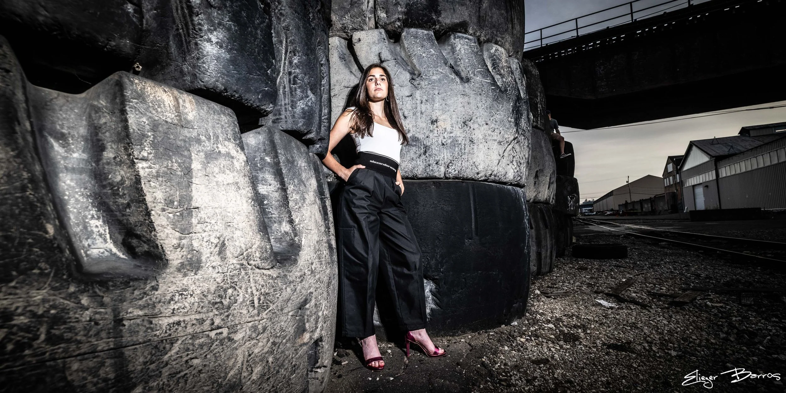 A woman standing in an industrial area with large black rocks behind her, wearing a white top, black pants and pink high heels.