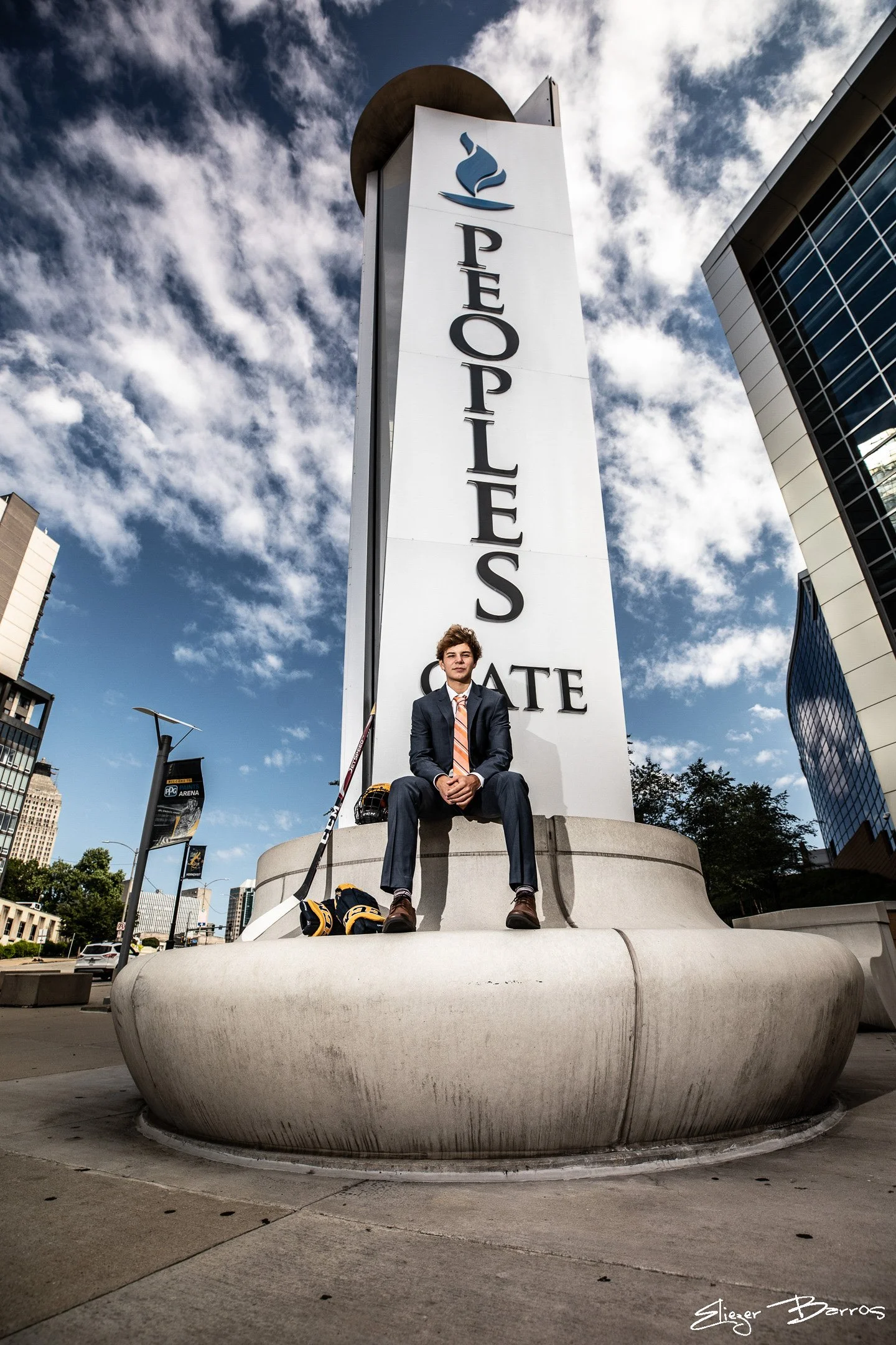 A young man in a business suit and orange tie sitting on a large, curved concrete fountain in front of a tall building with a sign that reads 'PEOPLE'S GATE' and a logo resembling a blue flame. The sky is partly cloudy and the cityscape includes othe