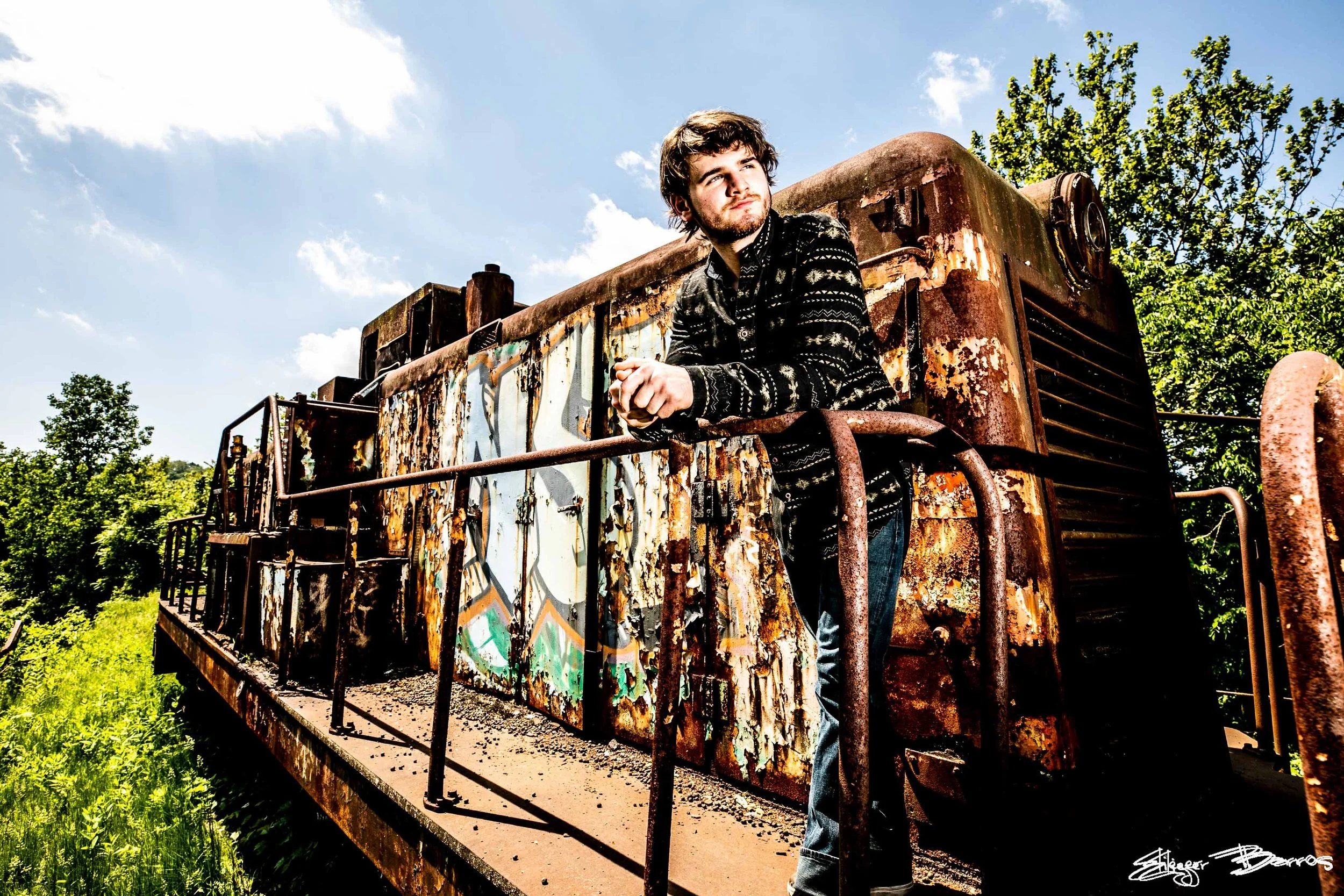 A young man with dark hair and a beard leaning on a rusted, graffiti-covered train car outdoors, with trees and a blue sky with clouds in the background.