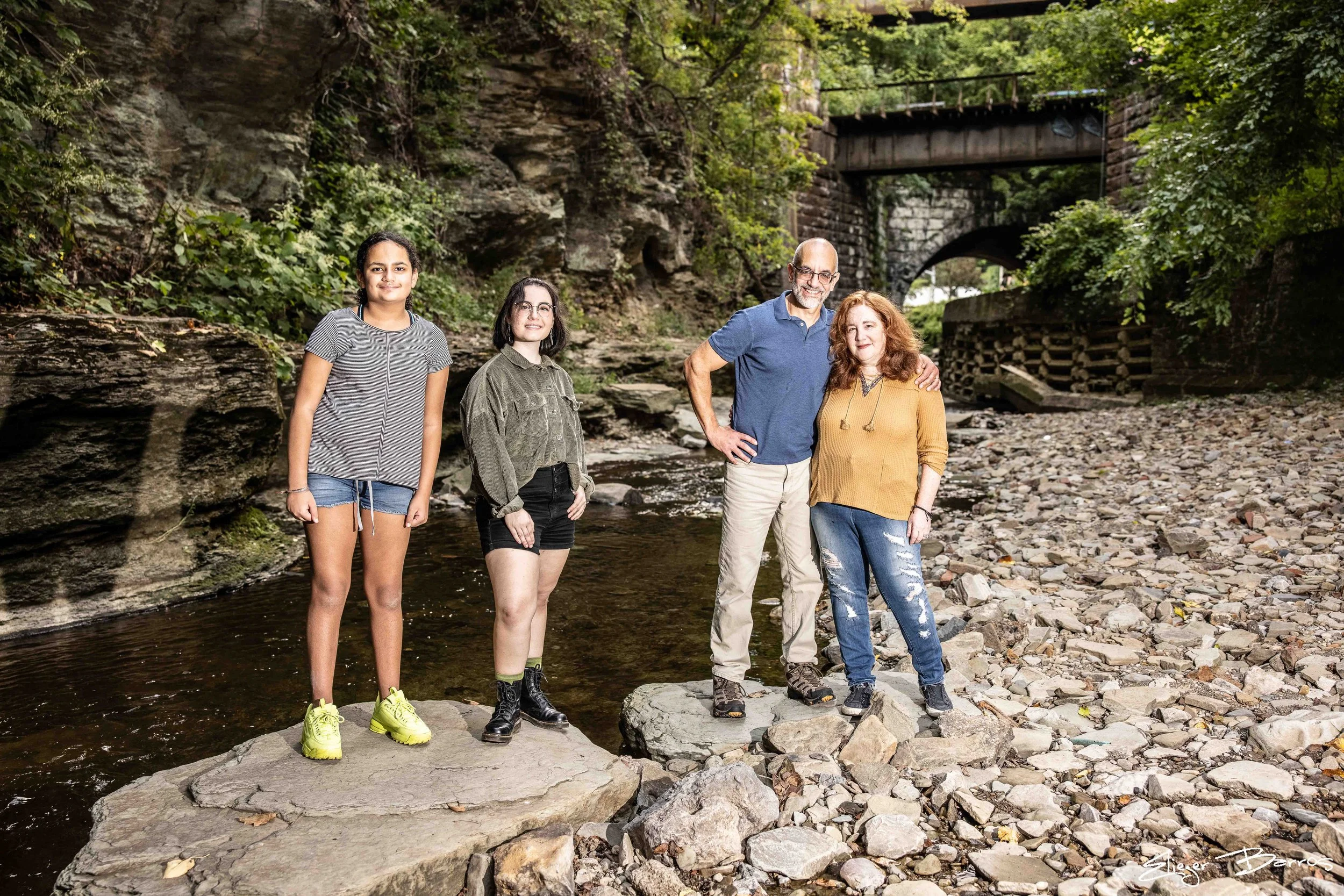 Four people standing on rocks in a creek with a stone bridge overhead, surrounded by trees and foliage.