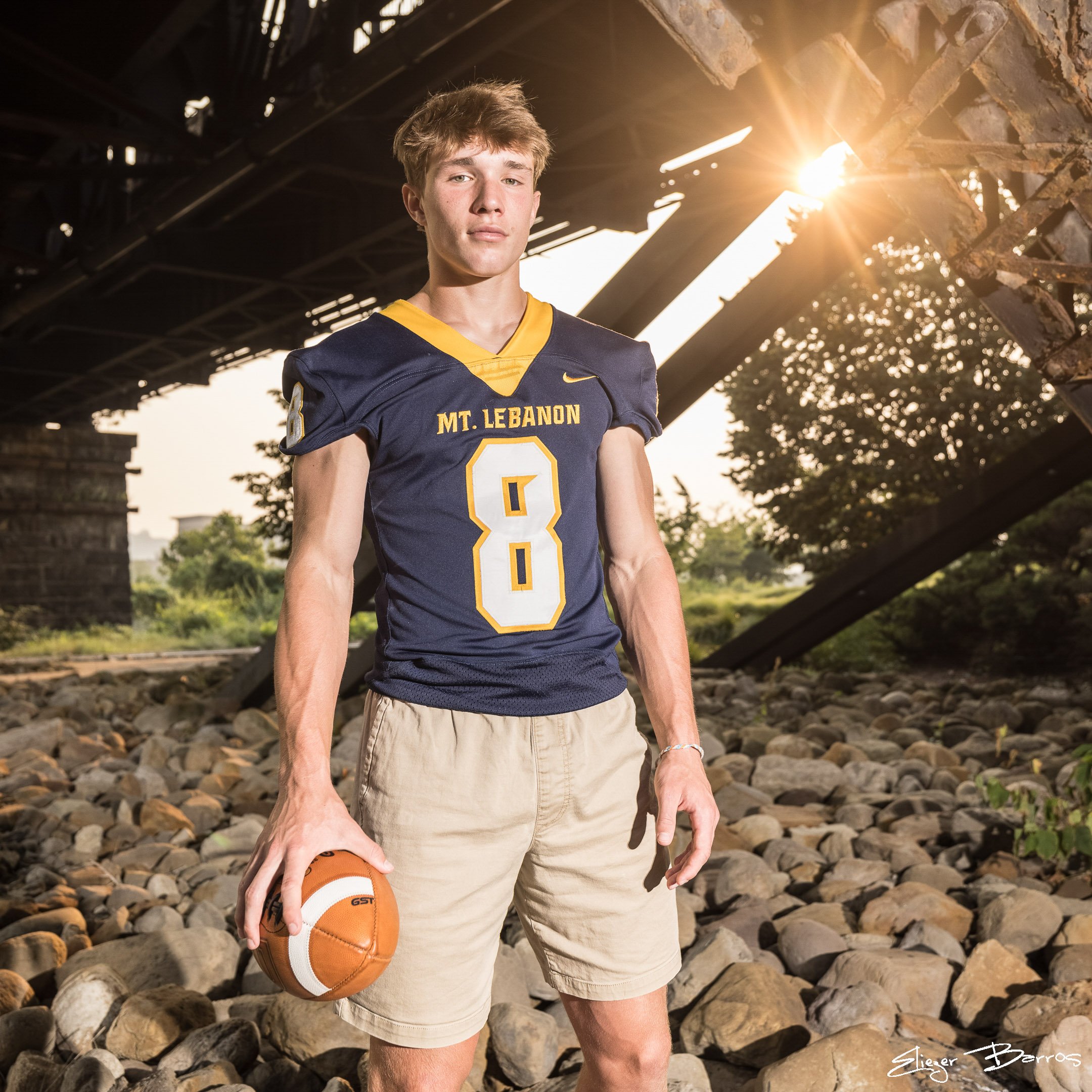 A young man in a sports jersey with "Mt. Lebanon" written on it, holding a basketball, standing outdoors on a rocky surface, with sunlight coming from behind and to the right.