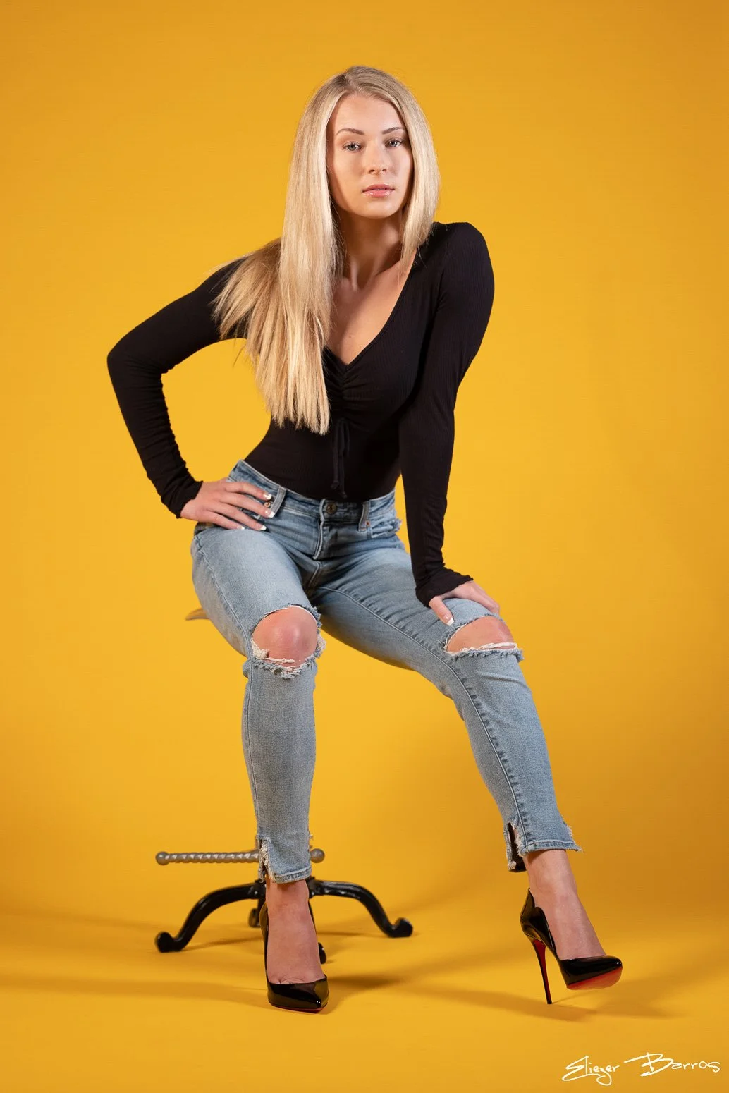 A blonde woman wearing a black long-sleeve top, ripped jeans, and high heels sitting on a stool against a yellow background.