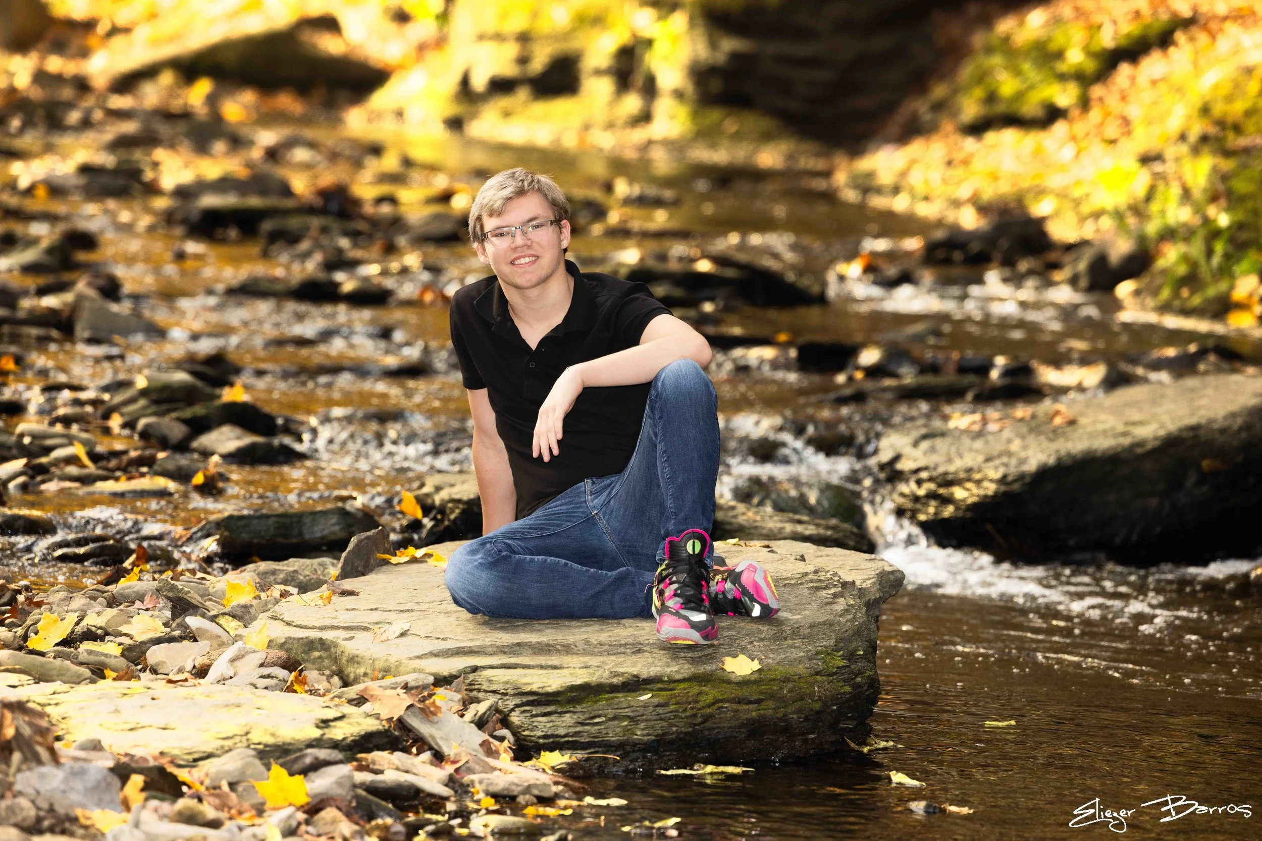 A young man with glasses, light-colored hair, wearing a black polo shirt and blue jeans, is sitting on a rock by a stream in a wooded area during autumn, with colorful fallen leaves around him.