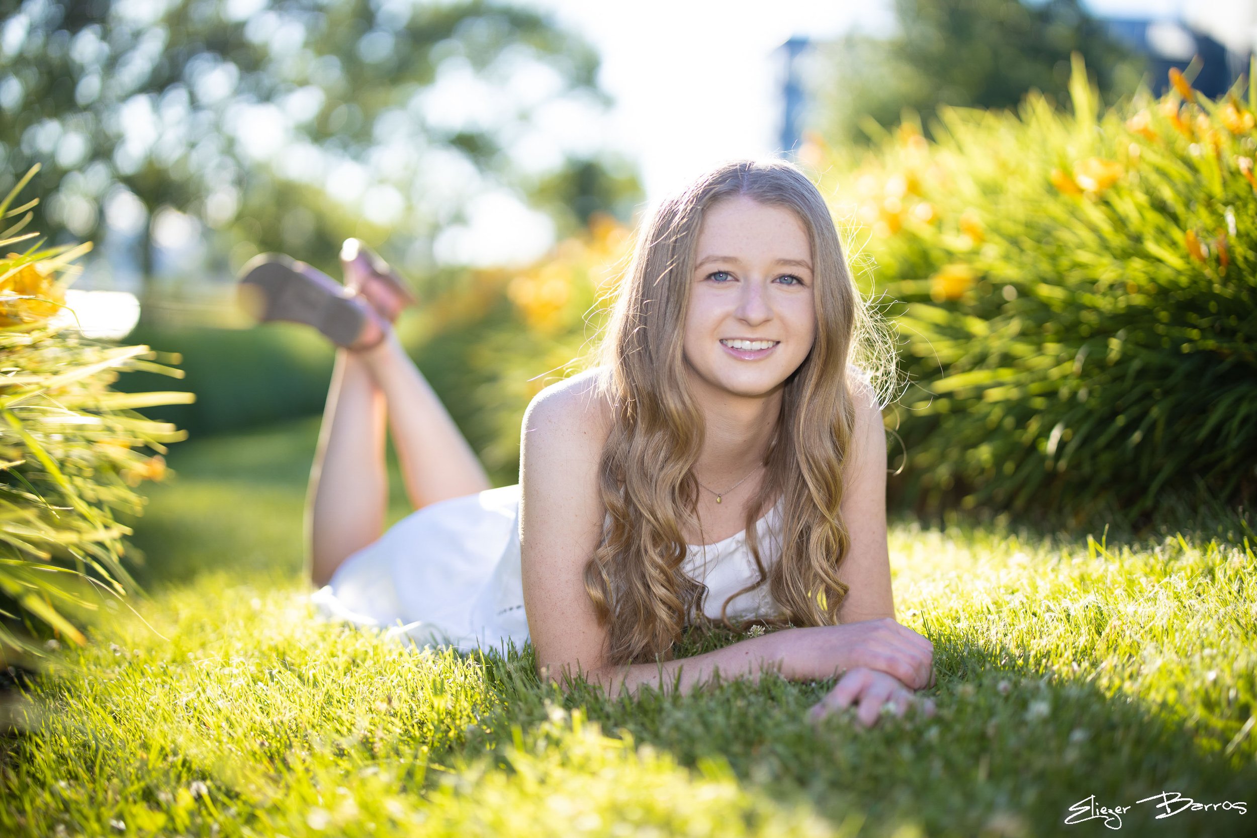 A young woman with long, wavy blonde hair lying on her stomach in a grassy yard, smiling at the camera with a bright outdoor background of blurred trees and bushes.