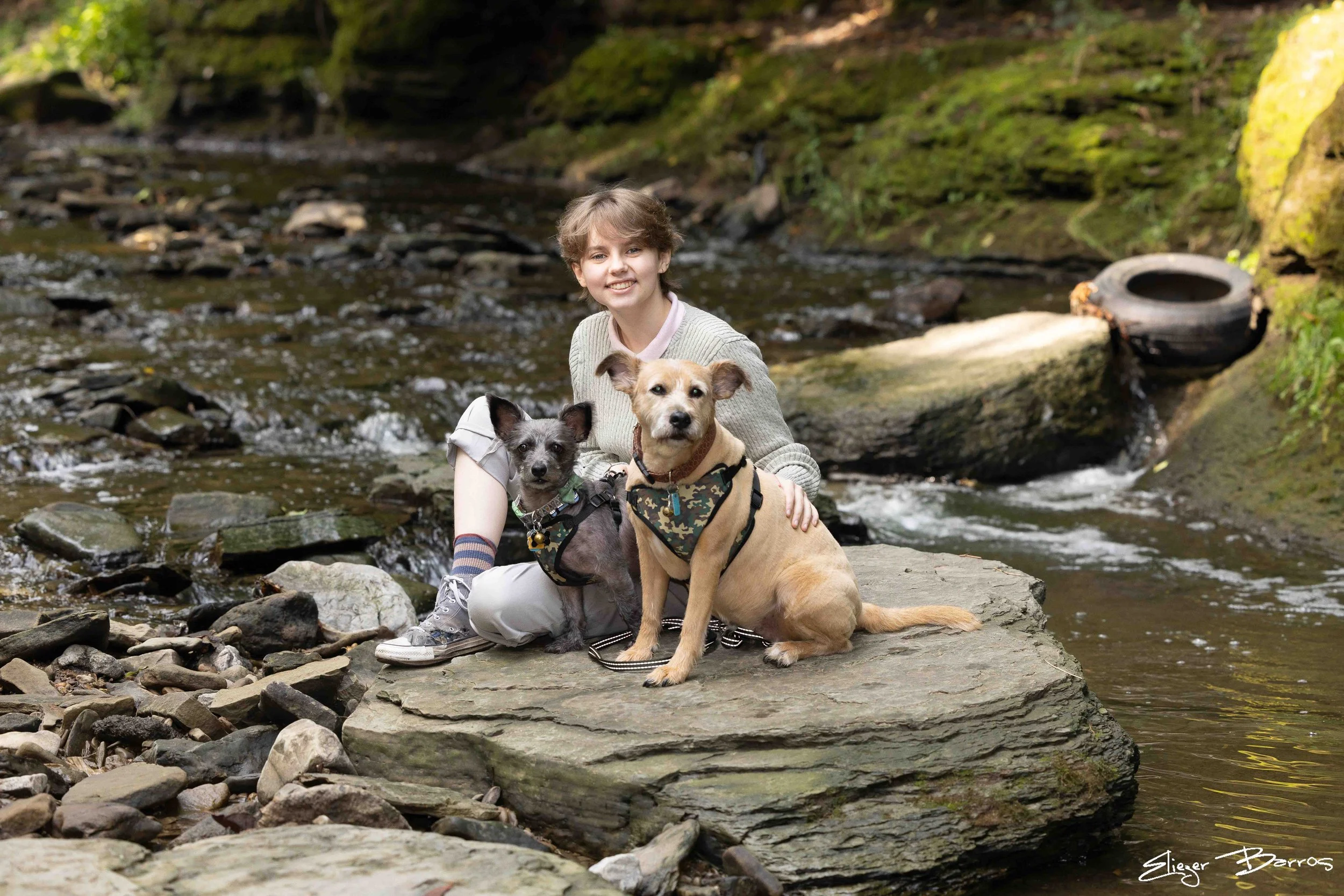 A young woman with short brown hair smiling while sitting on rocks by a stream, accompanied by two dogs, one tan with a camouflage vest and one smaller black and gray, in a forest setting.
