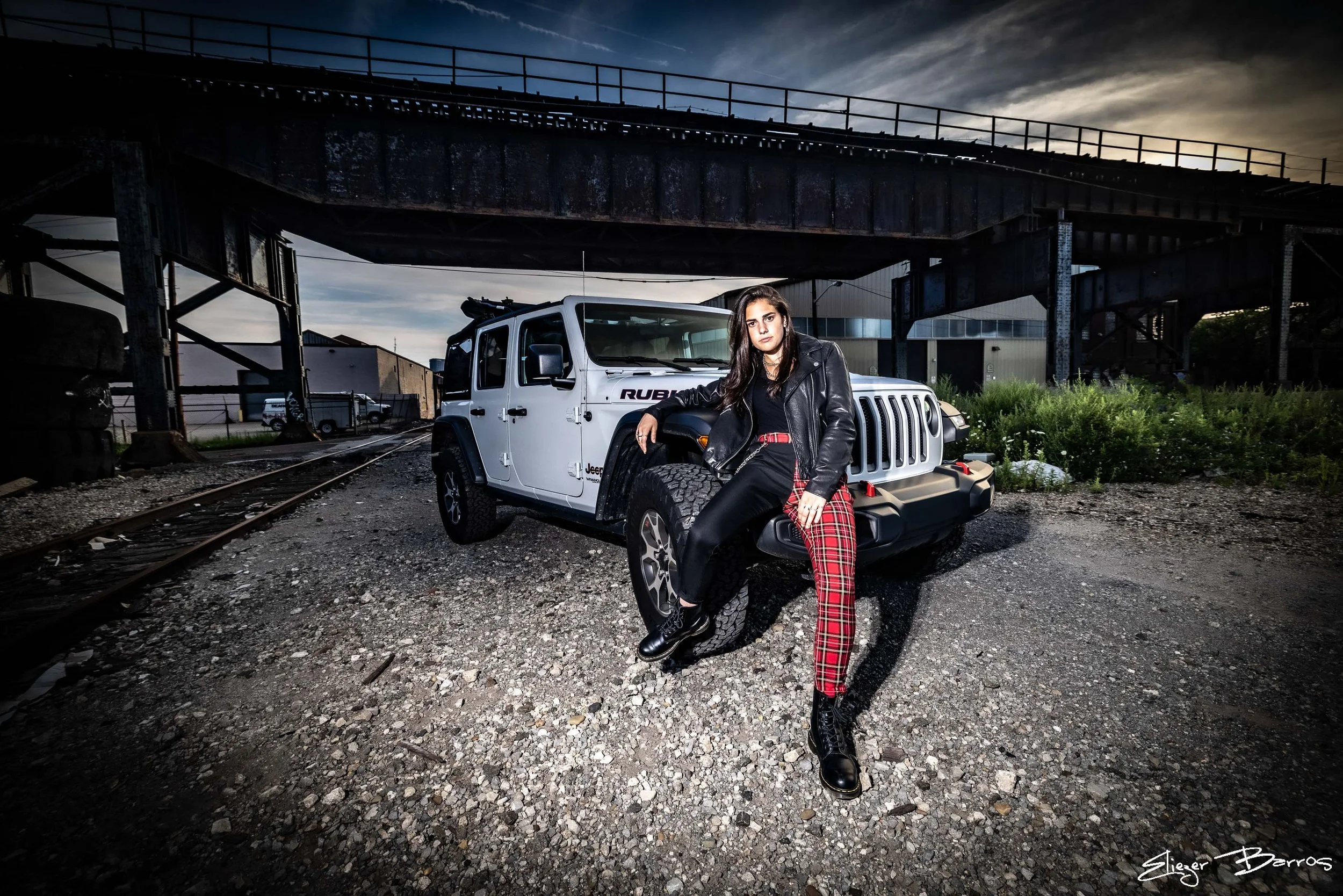 A young woman with dark hair and dark makeup leaning against a white Jeep Rubicon under an old bridge. She is wearing a black leather jacket, black shirt, red plaid pants, and black boots. The scene is outdoors, during dusk or early evening, with ind