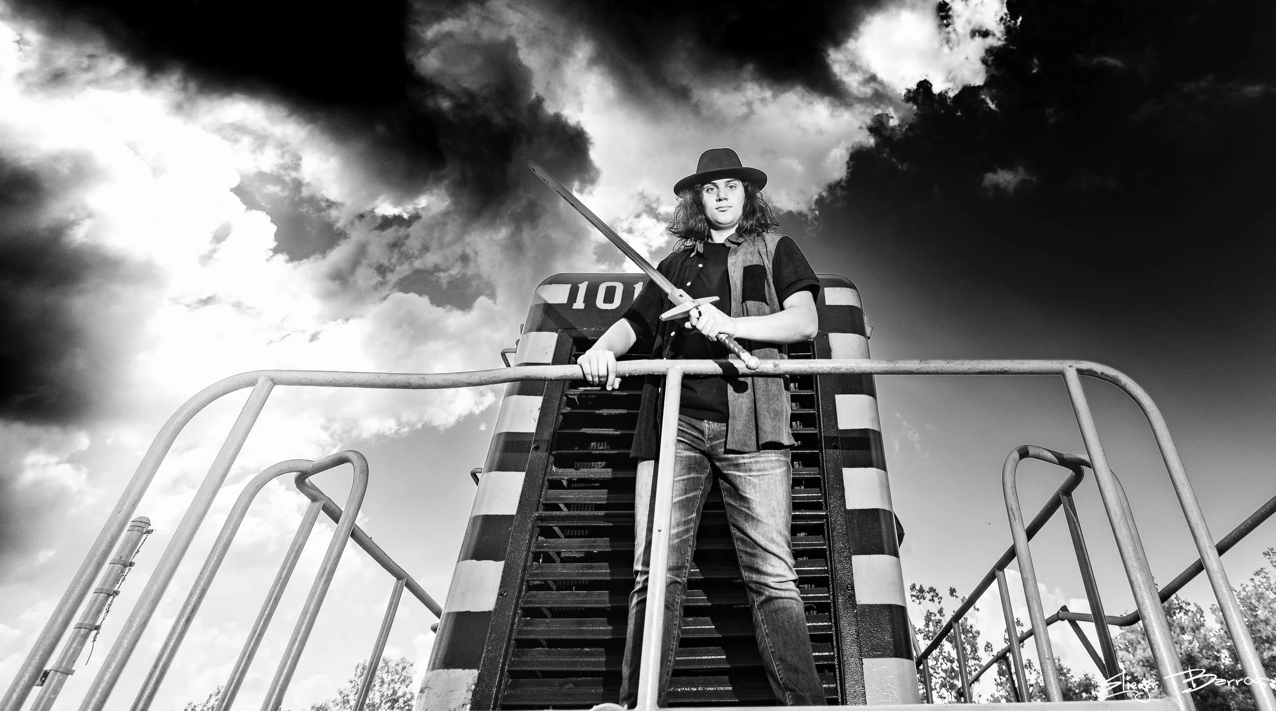 A young person with curly hair wearing a hat and a jacket, holding a sword, standing on top of a construction vehicle with striped barriers around it, against a cloudy sky.