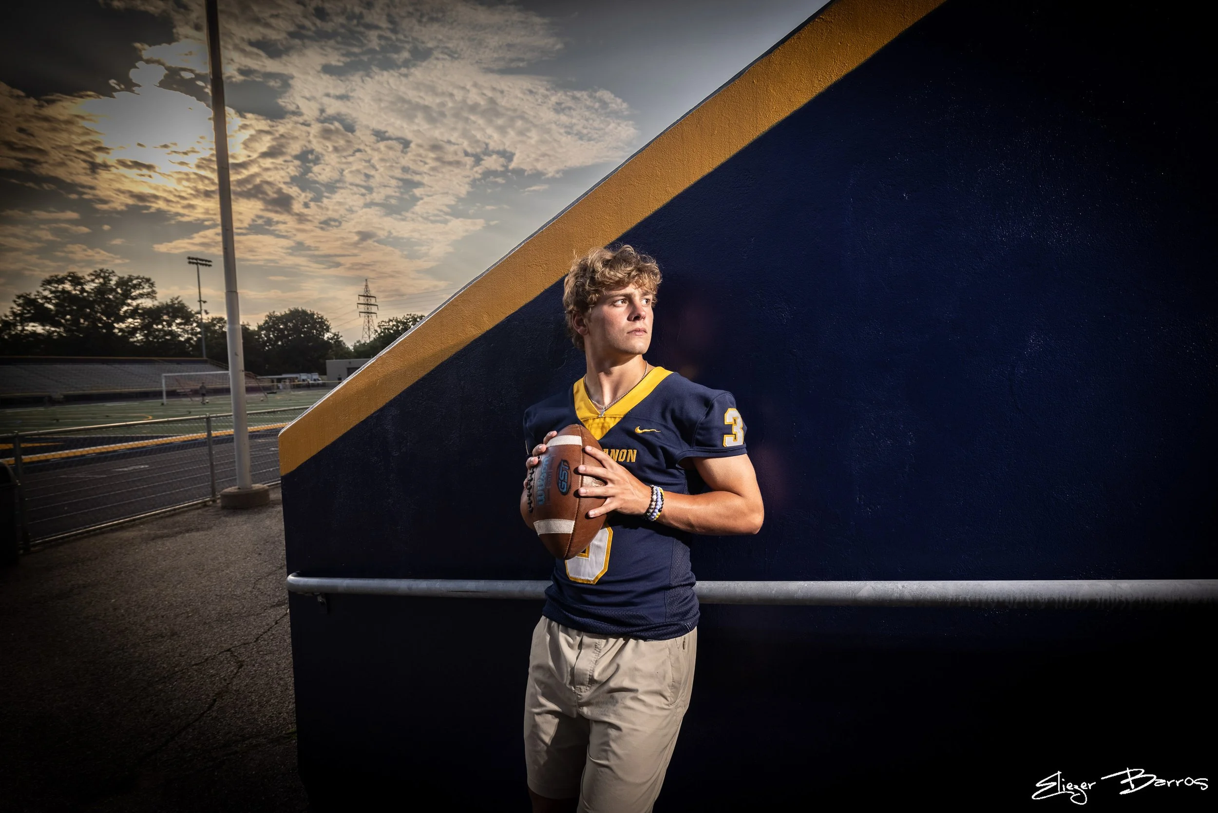 Senior Pictures Pittsburgh at high school stadium. Young man in blue and gold football jersey holding a football posing in front of school bleachers for athletic senior portraits.