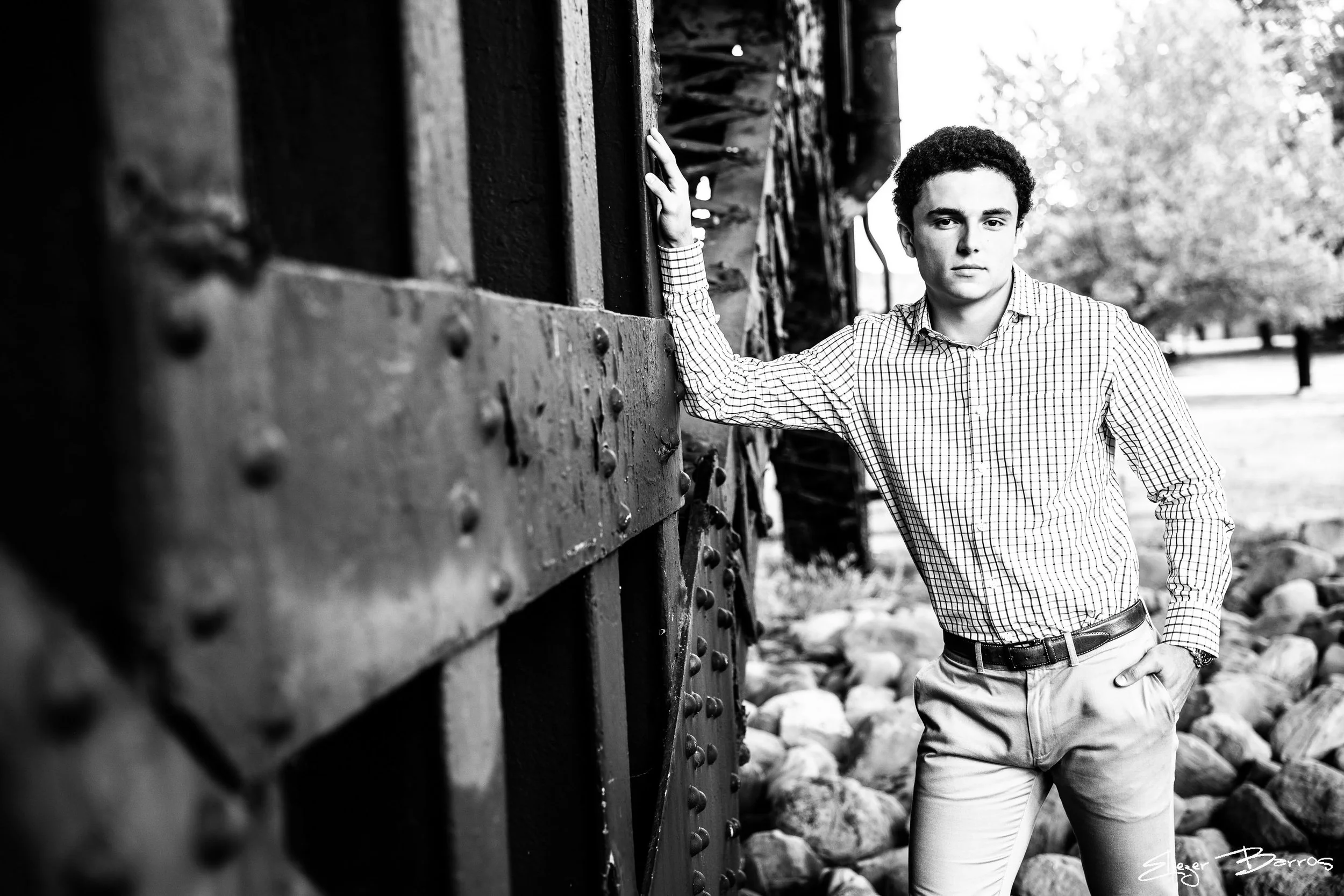 A young man with curly hair wearing a checkered shirt and light-colored pants standing outdoors near a train or railway structure.