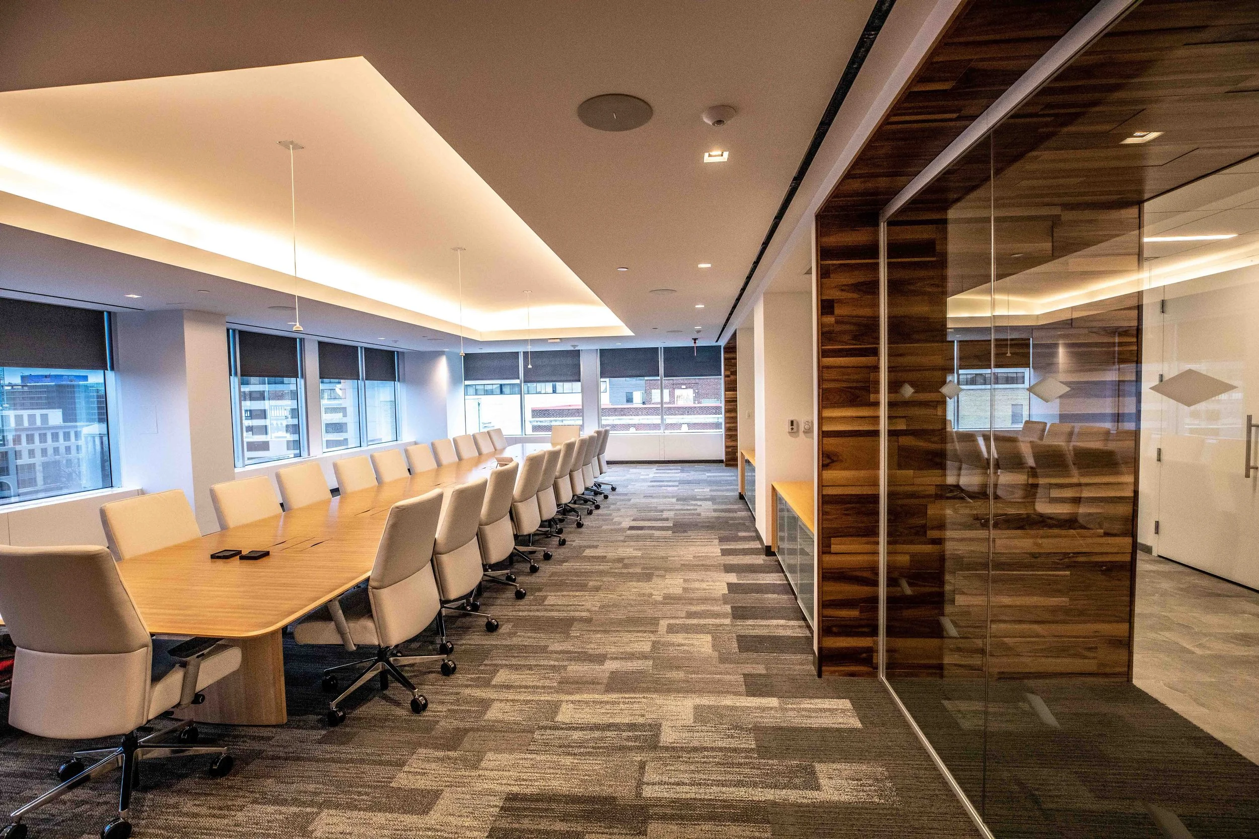Modern conference room with a long wooden table and beige chairs, windows showing cityscape, and a glass-walled area with wood paneling.