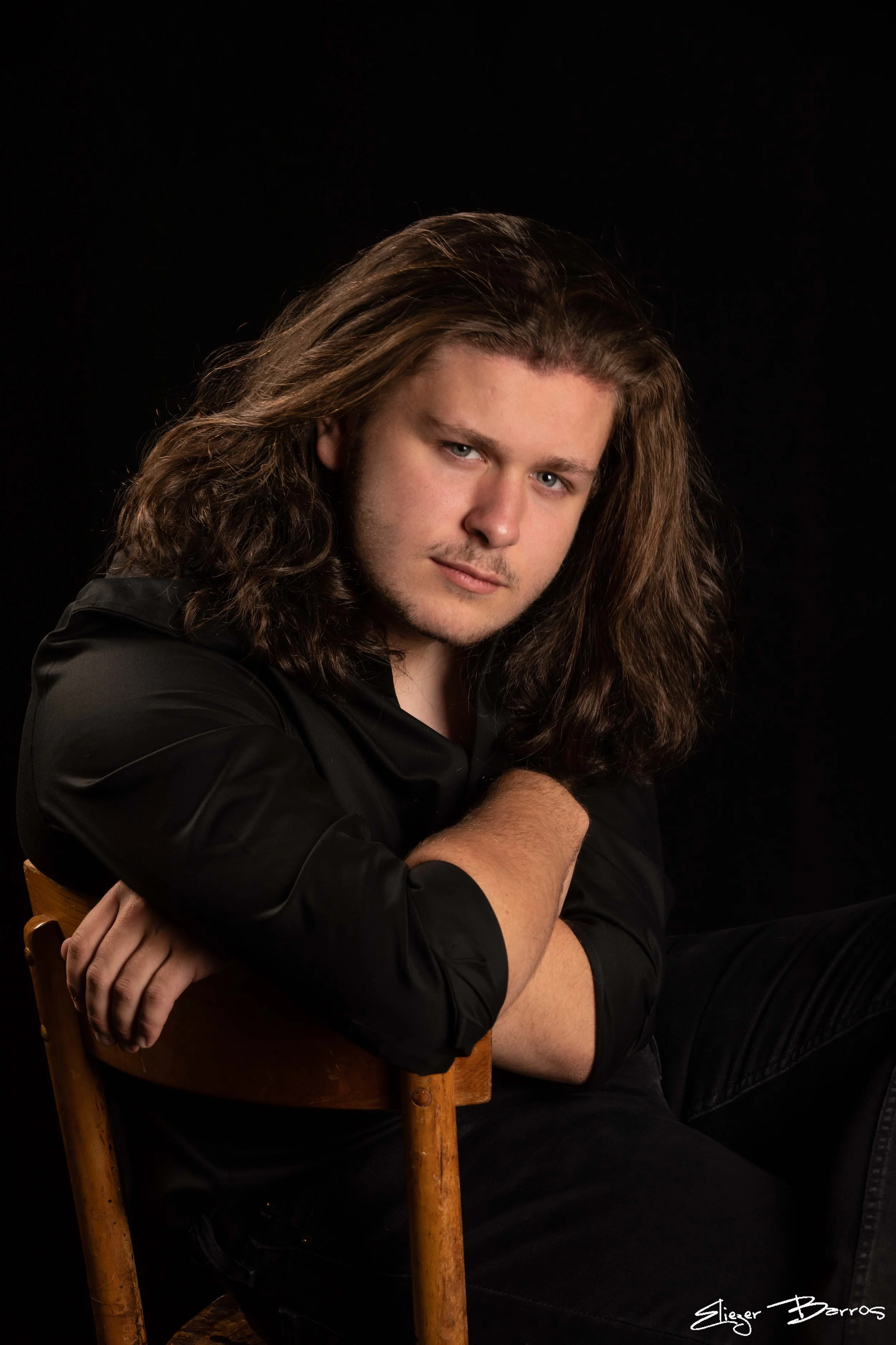 A portrait of a young man with long, wavy hair and blue eyes, wearing a black shirt, sitting with crossed arms on a wooden chair against a black background.