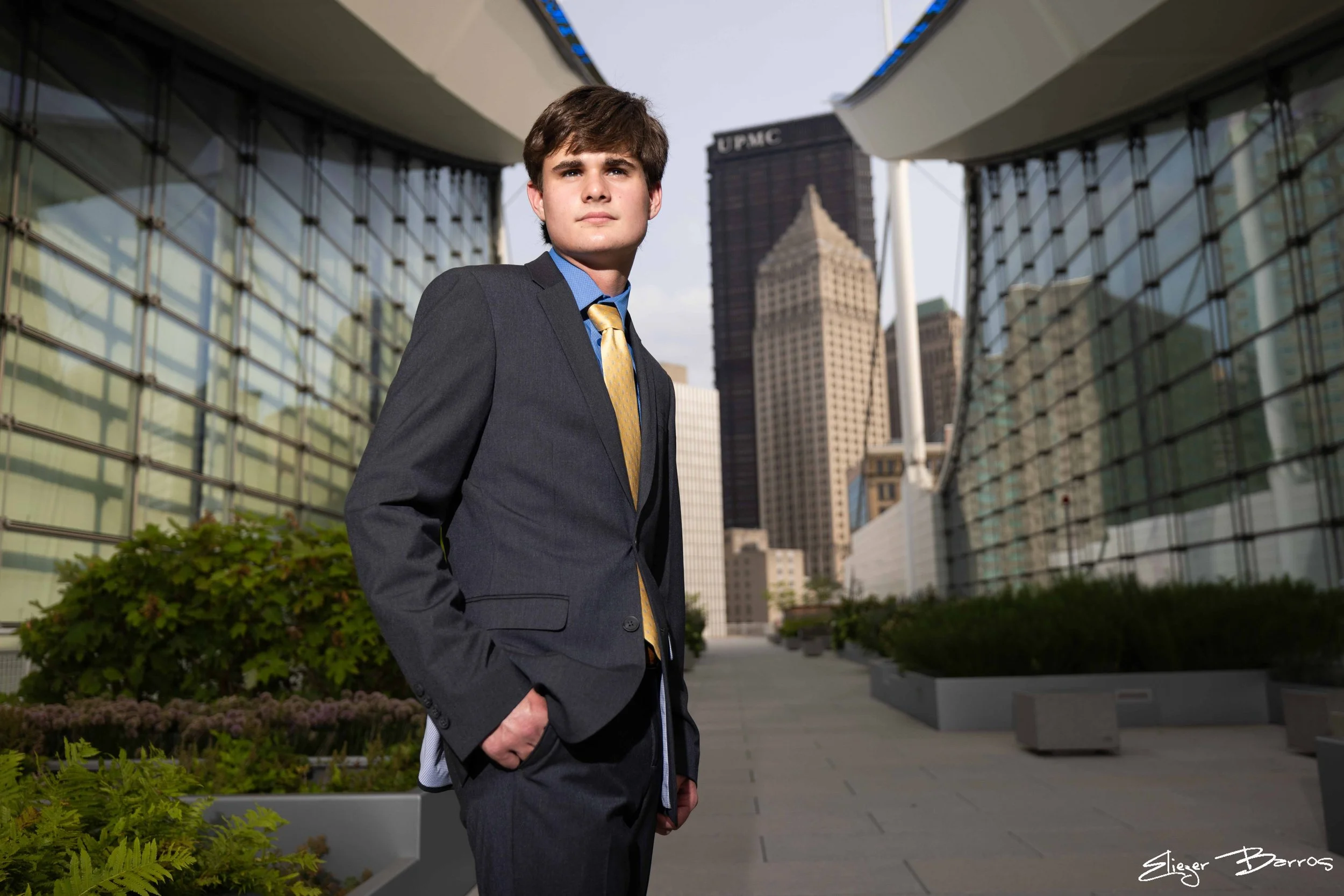 Senior Pictures Pittsburgh Downtown. Young man in a gray suit and gold tie posing with hands in pockets in front of modern glass skyscrapers for stylish urban senior portraits.