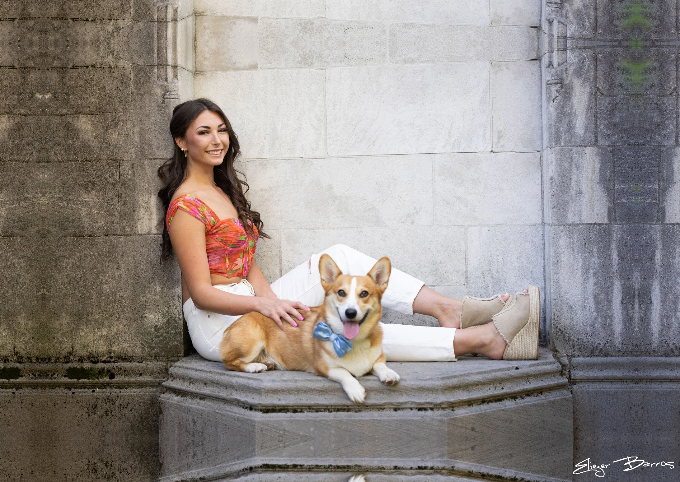 A young woman with dark hair in a colorful top and white pants sitting on steps against a stone wall, smiling, with a cute corgi dog with a blue bow sitting nearby.