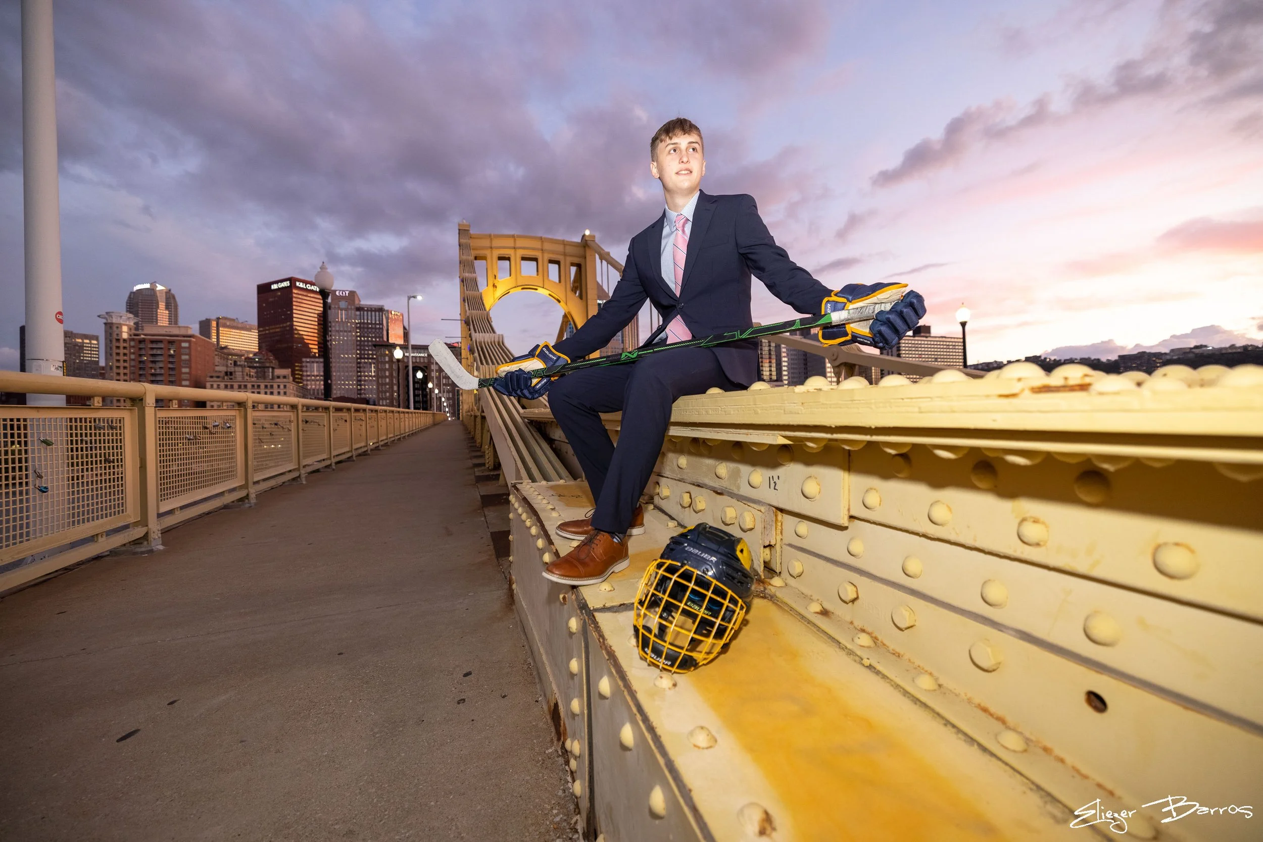 Young man in business suit sitting on a yellow bridge railing at sunset, holding a hockey stick, with a hockey helmet and gloves nearby, city skyline in the background.