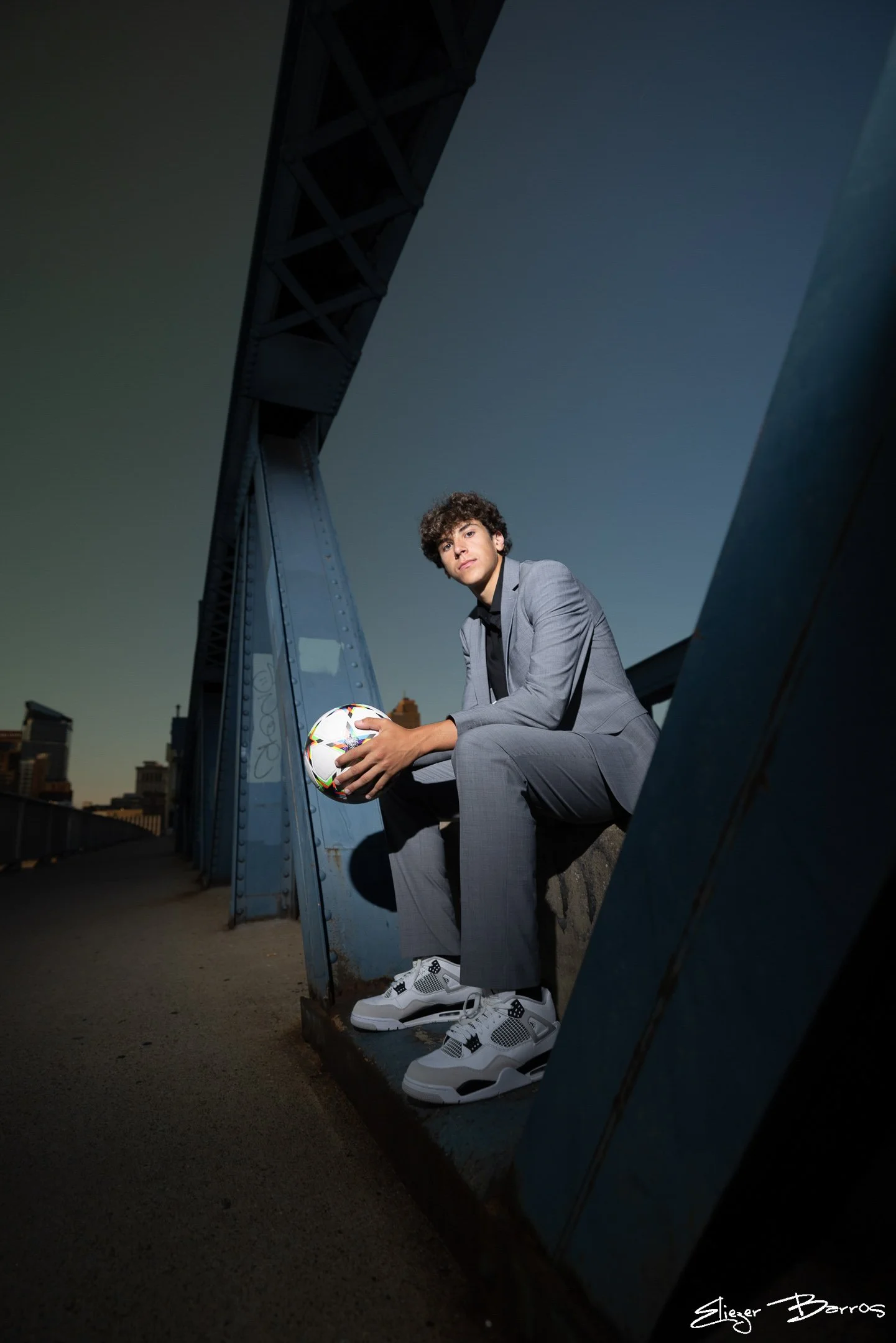 A young man dressed in a gray suit, white sneakers, and white and black socks, sitting on a ledge under a bridge during dusk, holding a colorful soccer ball with city buildings in the background.