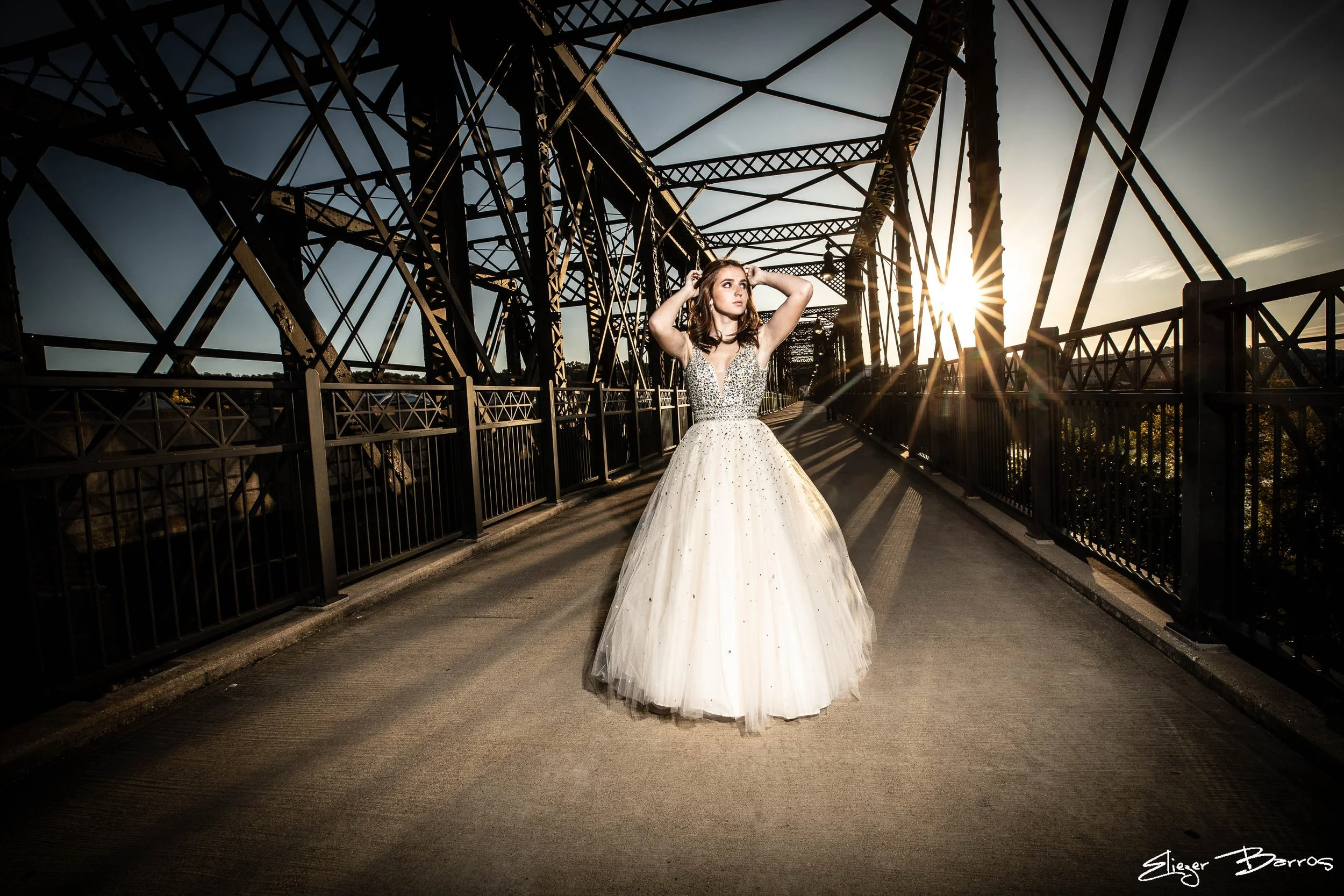 Senior Pictures Pittsburgh at Hot Metal Bridge. Young woman in a sparkling white ball gown posing on the historic steel bridge with dramatic sunset light flare and river views for elegant industrial senior portraits.