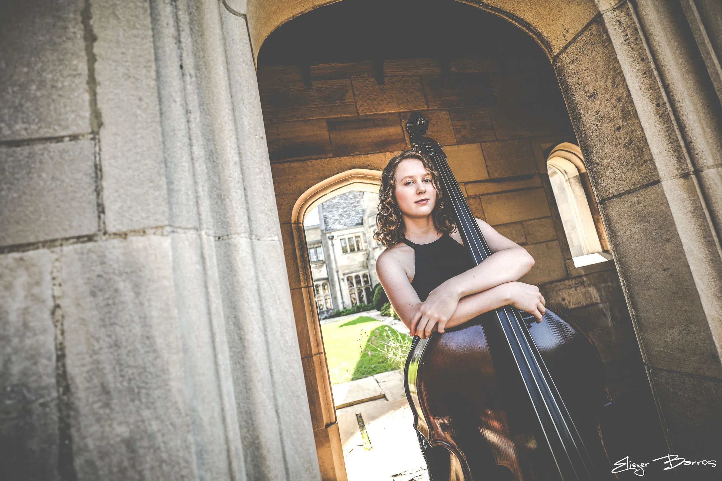 A young woman with curly hair holding a double bass in an archway of an old stone building, with a courtyard and windows in the background.