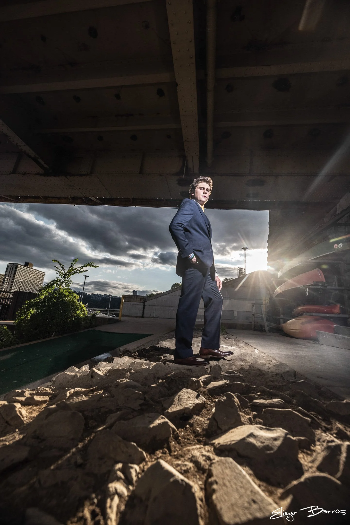 A man in a dark suit and brown shoes standing outdoors under a bridge, with a cloudy sky and cityscape in the background, and sun rays shining behind him.