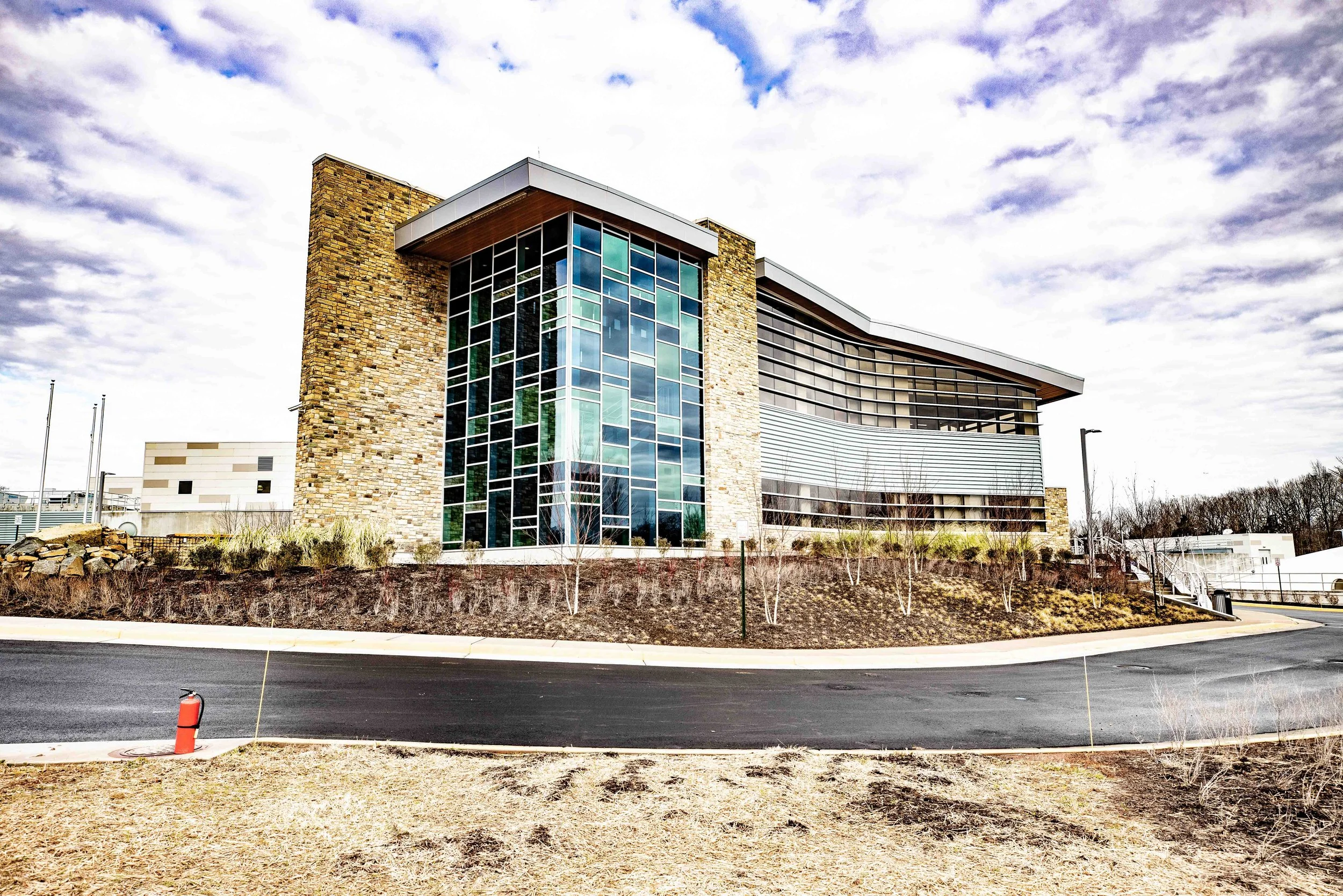 A modern building with large glass windows and stone walls under a cloudy sky, situated in a landscaped area with young trees and a paved road in the foreground.