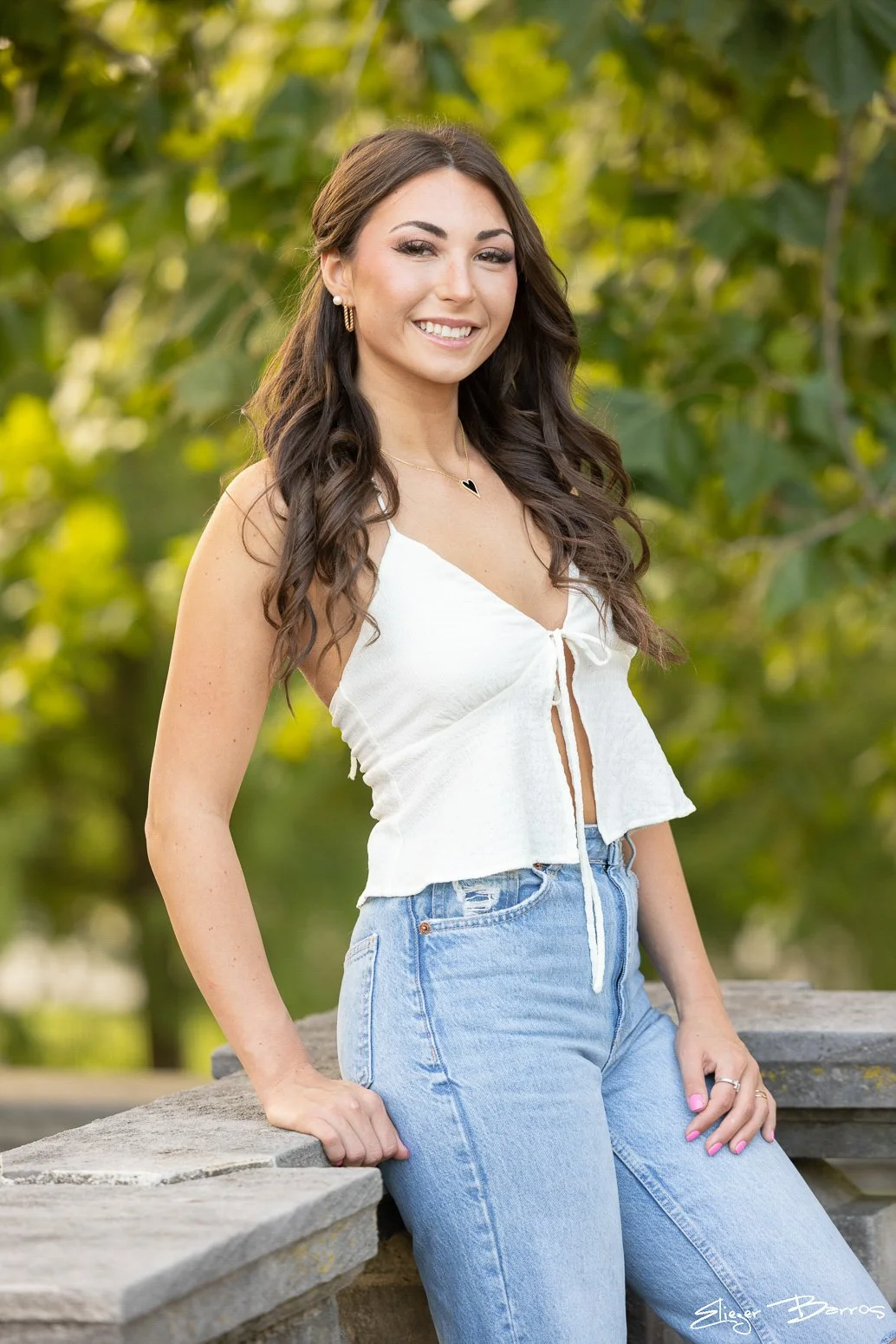 A young woman with long wavy brown hair smiling outdoors, wearing a white sleeveless top and light blue jeans, standing near a stone ledge with green trees in the background.