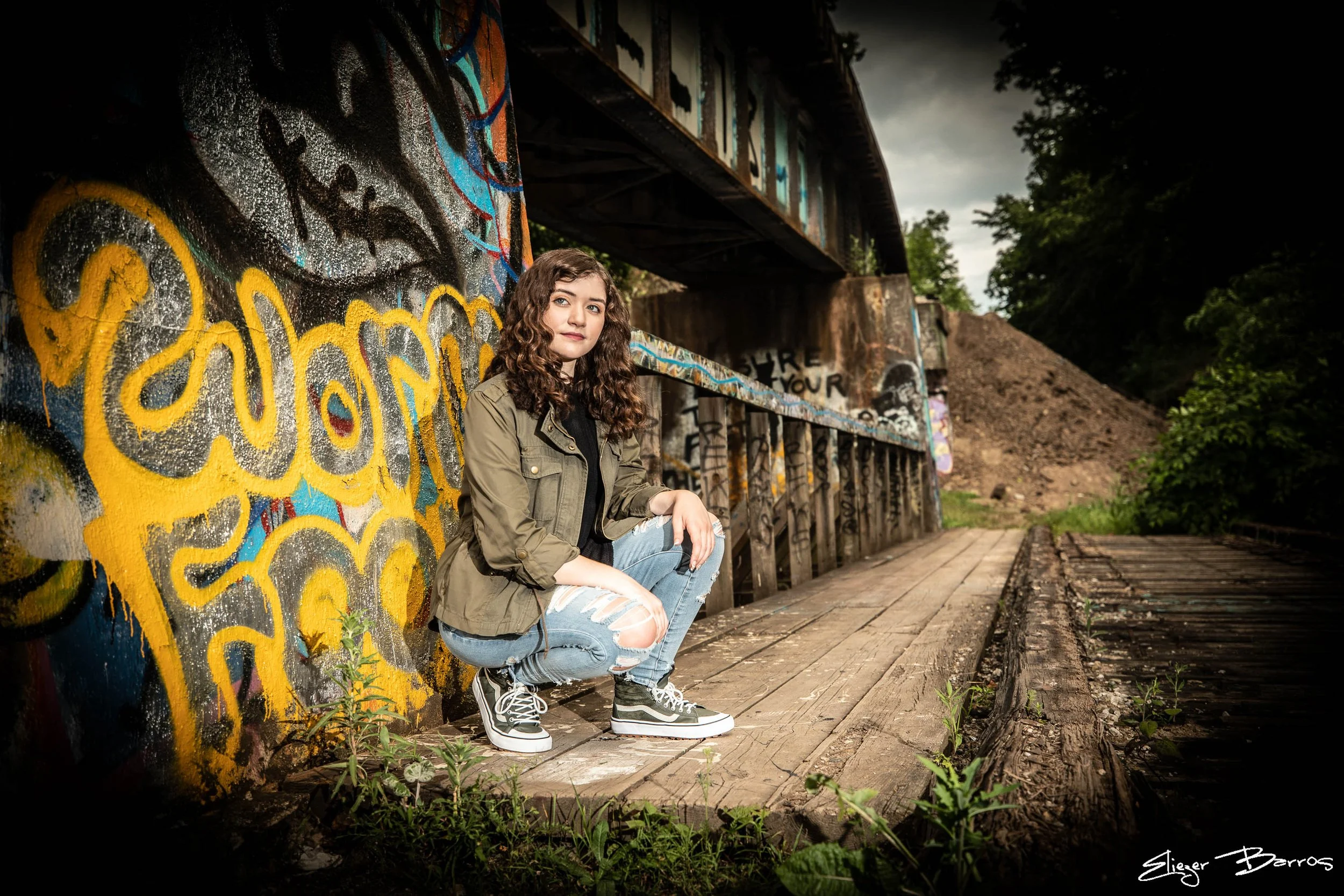 A young woman with curly hair, wearing a green jacket, ripped jeans, and black and white sneakers, crouching in front of a graffitied wall underneath an old bridge in a forested area.