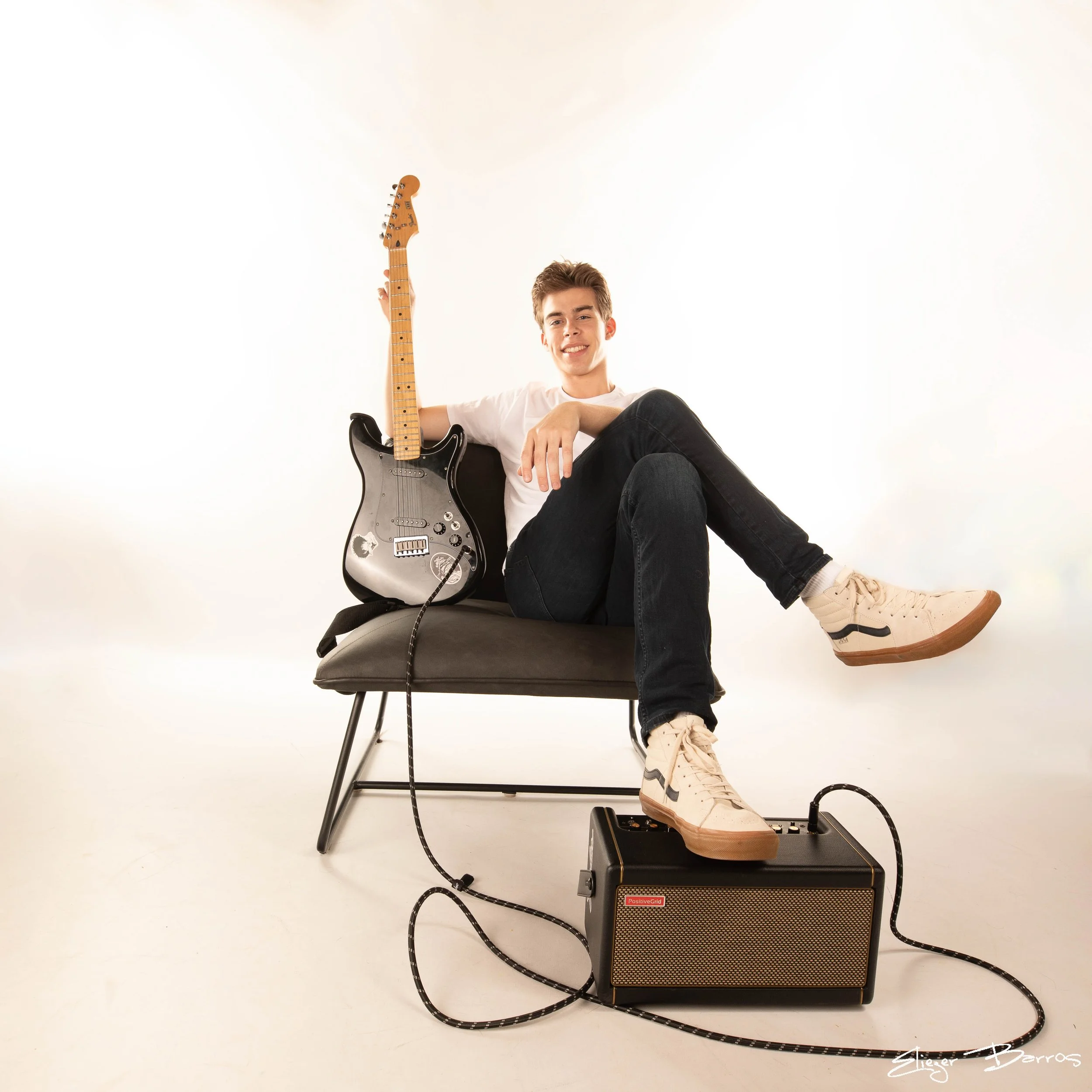 A young man sitting on a black chair with a guitar resting on his shoulder and smiling at the camera. He is wearing a white t-shirt, black pants, and beige sneakers. In front of him, there is a black amplifier with a foot pedal connected by cables. T