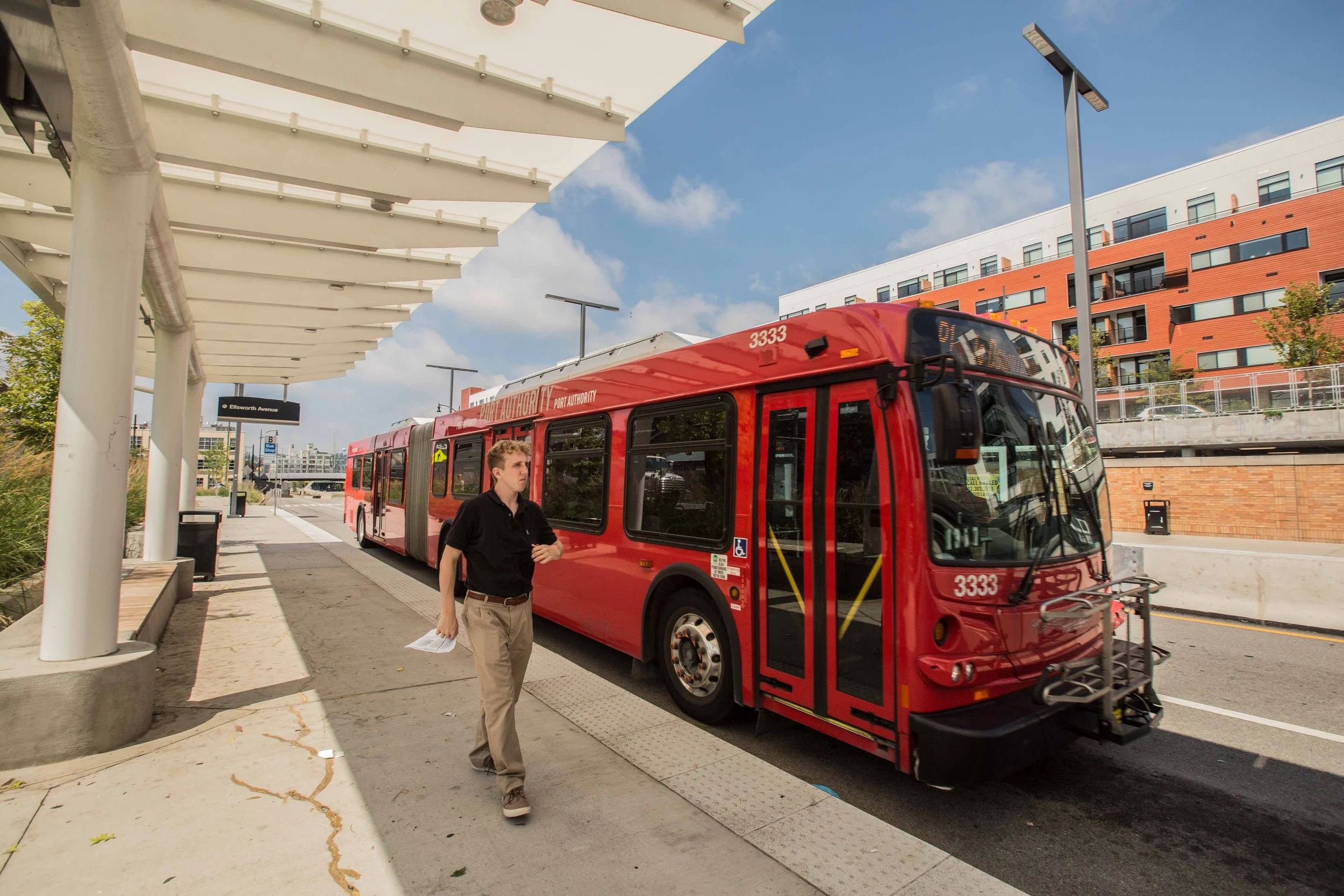 A red electric bus at a bus stop on a city street. A man is walking nearby holding papers, with a modern building and blue sky in the background.