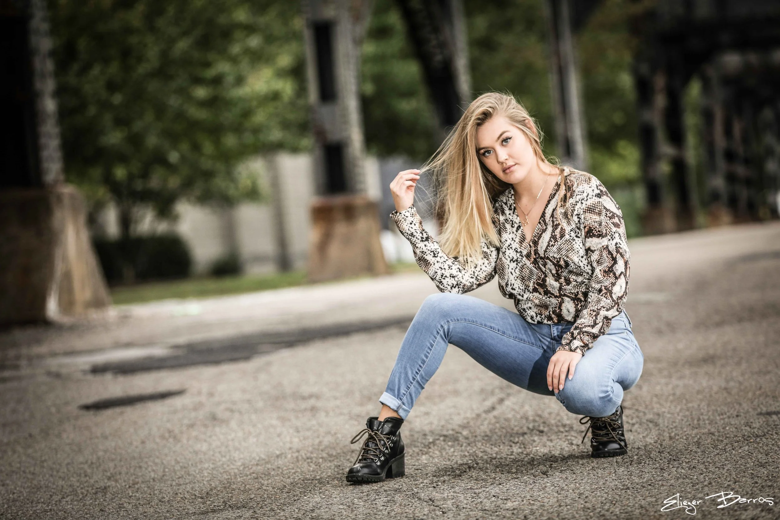 A young woman with long blonde hair wearing a snake print blouse, jeans, and black combat boots, squatting on the street with green trees and a building in the background.