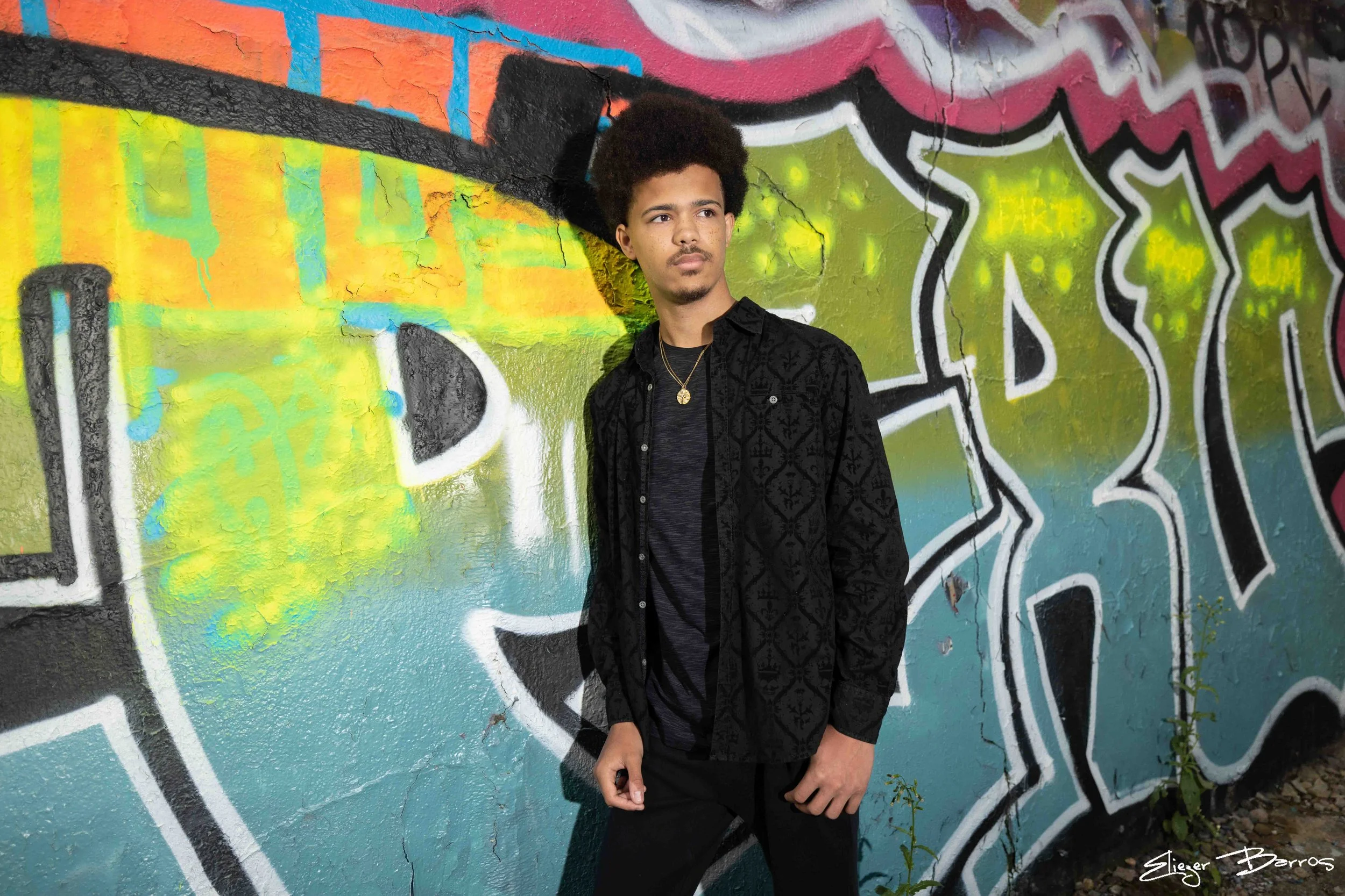 A young man with curly hair standing in front of a colorful graffiti wall.