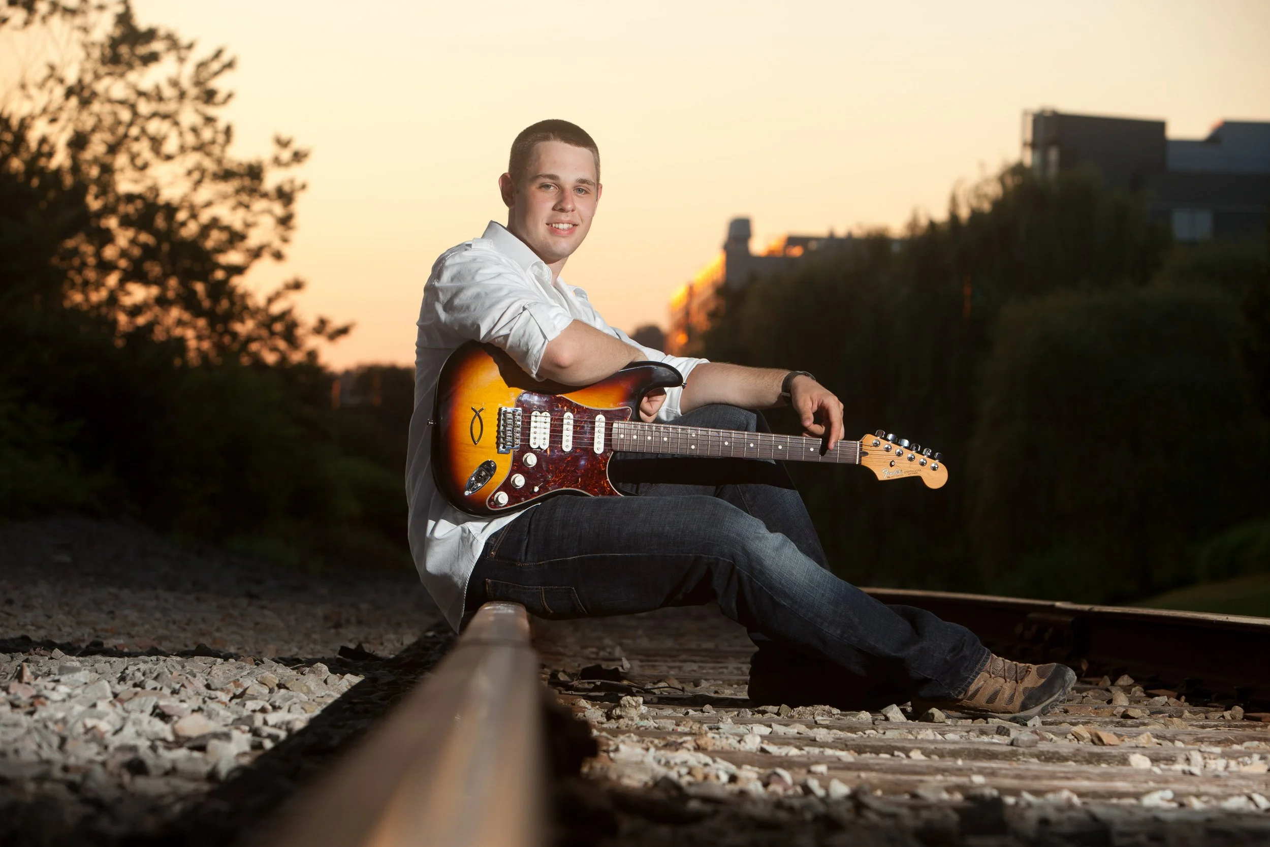 Young man sitting on railroad tracks, holding an electric guitar, looking at the camera with a sunset in the background.