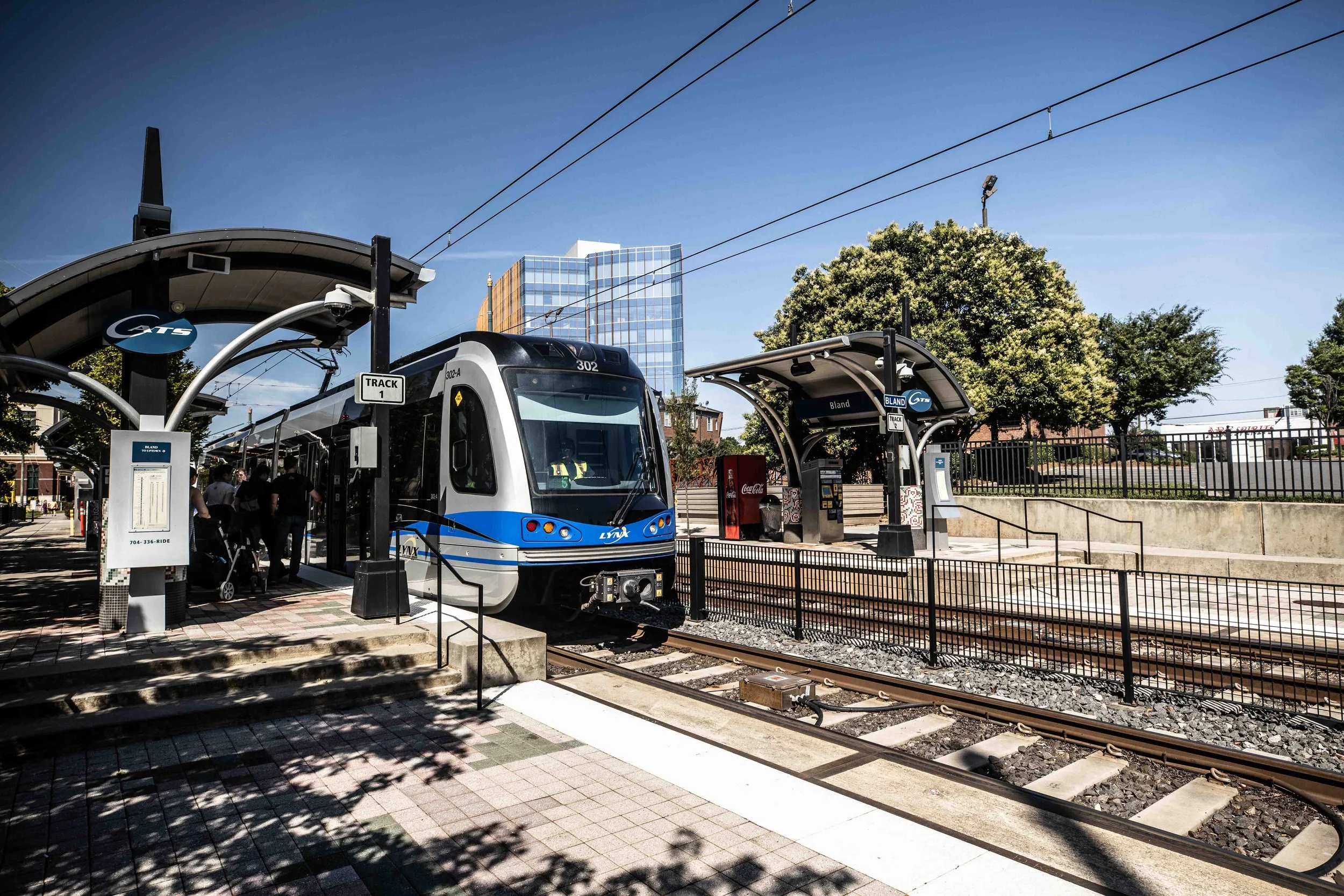 A modern light rail train at a station with a curved canopy, people waiting, and a cityscape with tall buildings and trees in the background.