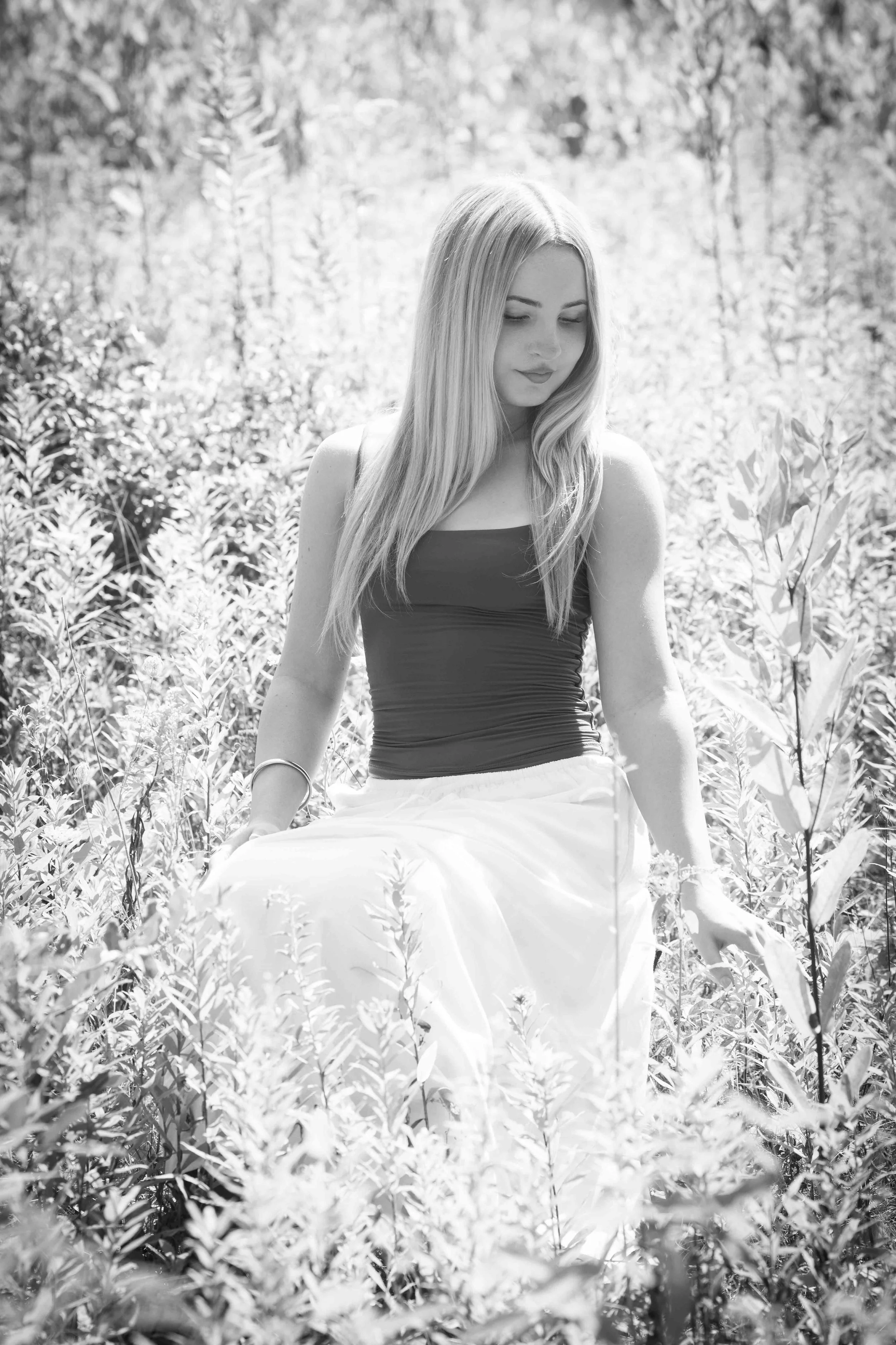 A young woman with long hair sitting outdoors among tall plants, with a blurred background of trees and foliage in black and white.