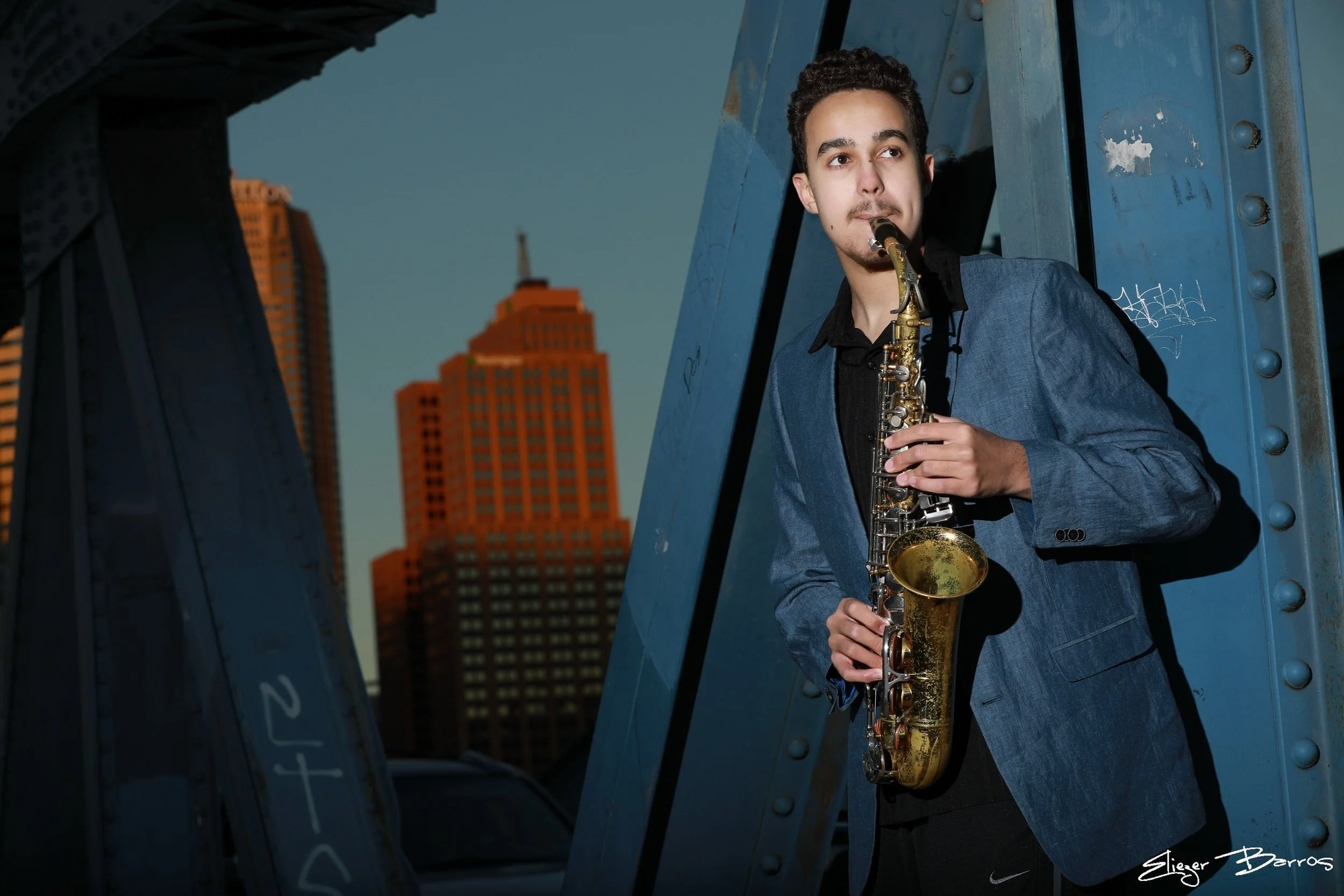A young man wearing a blue blazer playing the saxophone in an urban setting with tall buildings in the background.