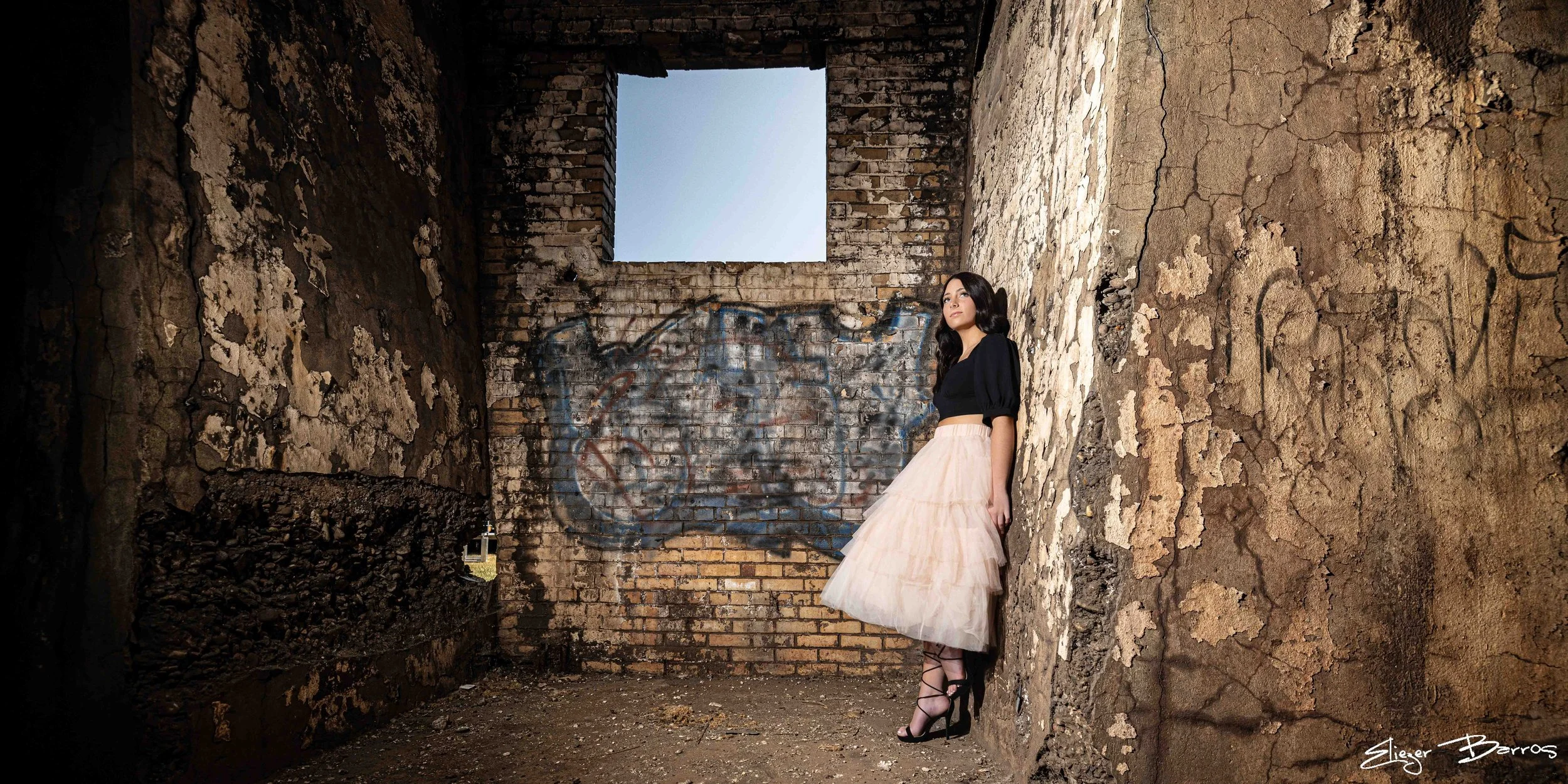 A woman in a black top and long light pink skirt standing in an abandoned, graffitied brick building with open walls and a window showing the sky.